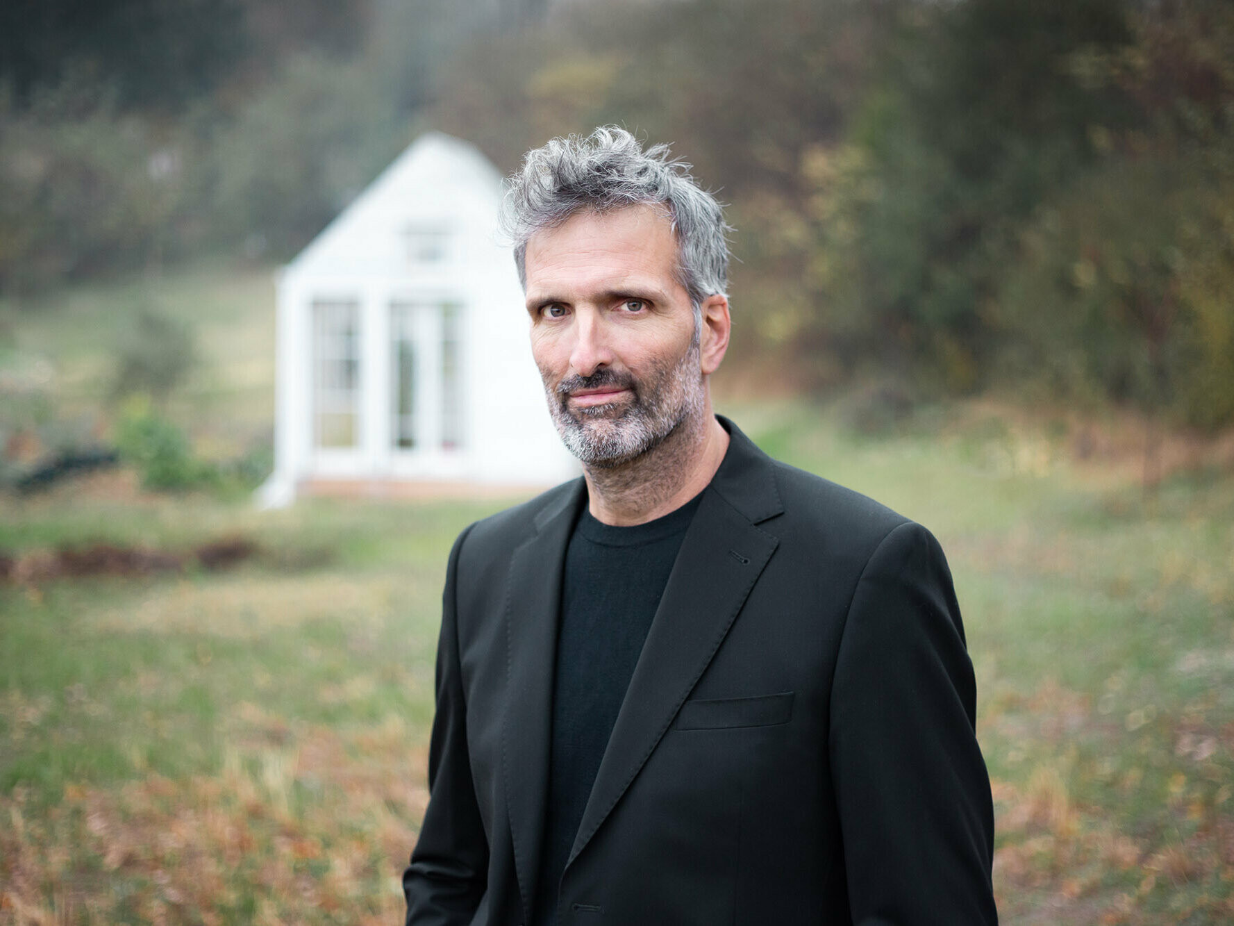 Portrait of the lead architect Carlo Pierantozzi from zooform architecture + design, behind him a meadow with a forest behind it and a small white building can be seen out of focus.
