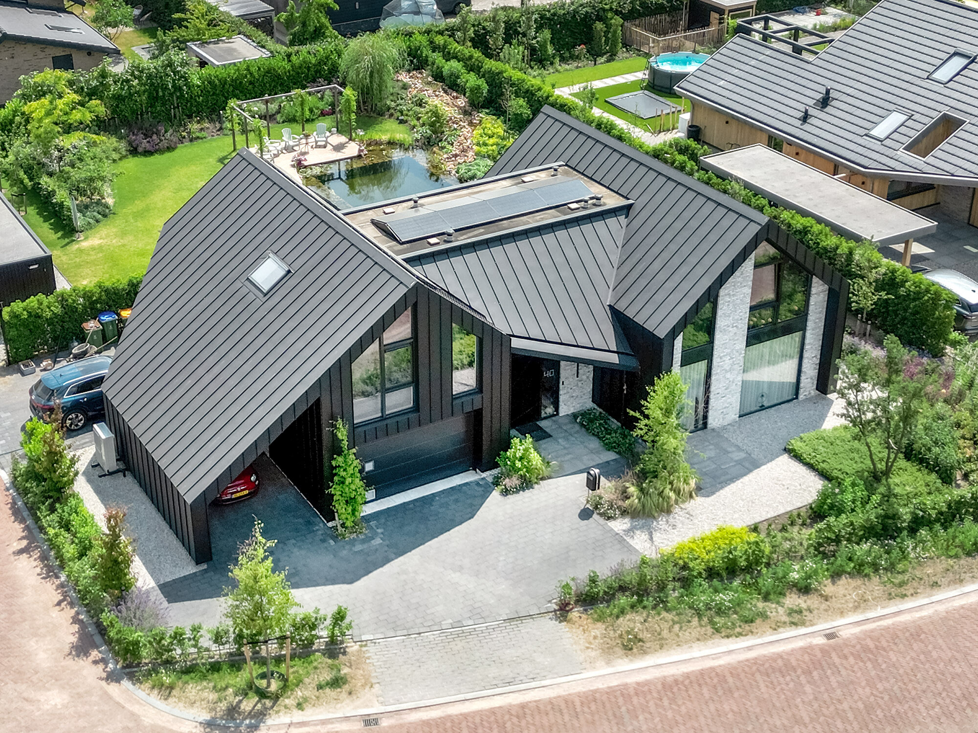 Aerial view of a modern detached house with a steep, black Prefalz pitched roof and integrated solar modules. The architecture combines large glass fronts and geometric shapes. A carport is integrated into the building. The property has a well-tended garden with a pond and terrace, surrounded by a quiet, suburban neighbourhood.