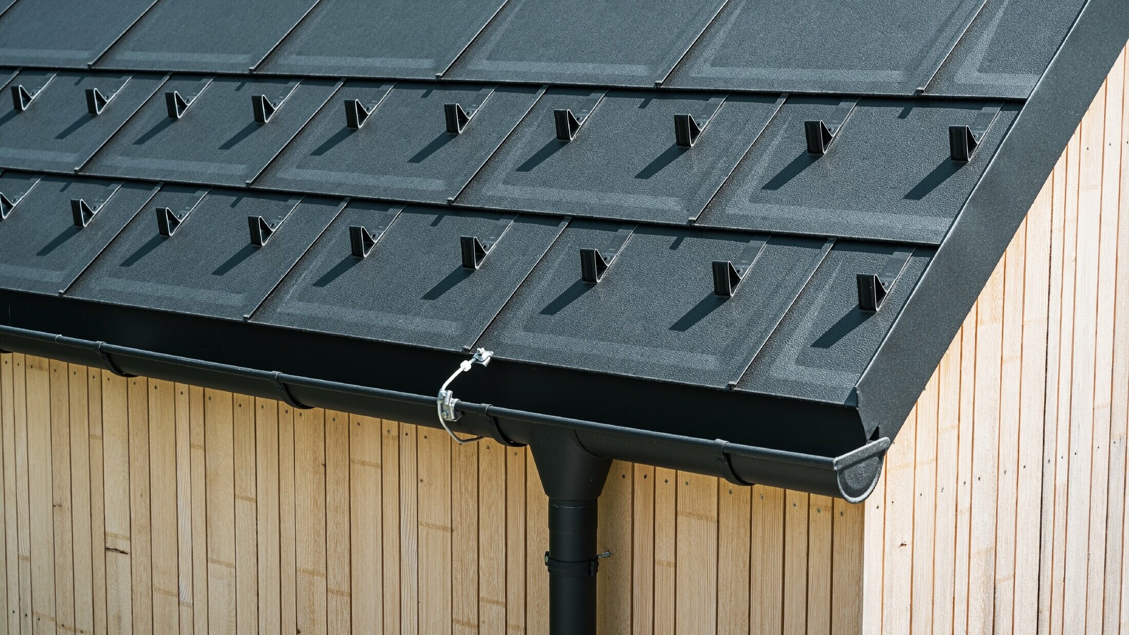 Close-up of PREFA roof tile R.16 with snow guards, gutter and downpipe in black P.10 incl. lightning protection and a vertical wooden façade