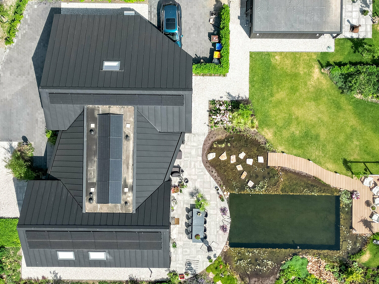 Aerial view of a modern detached house with a complex roof system made of black Prefalz, solar panels and several dormers. The property has a well-kept garden with lawns, a large pond, a wooden footbridge and a seating area. Paved paths and a driveway complete the picture of the neatly landscaped grounds surrounding the house.