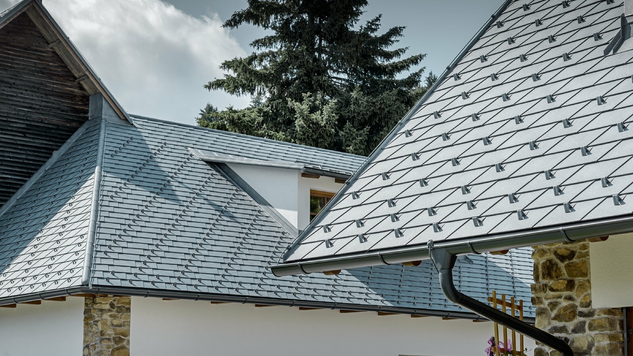 Close up of a PREFA aluminium roof cover; roof shingles in stone grey with the PREFA aluminium gutter in anthracite; an eyebrow dormer with standing seam roof can be seen in the background. The façade is white with integrated stone elements.
