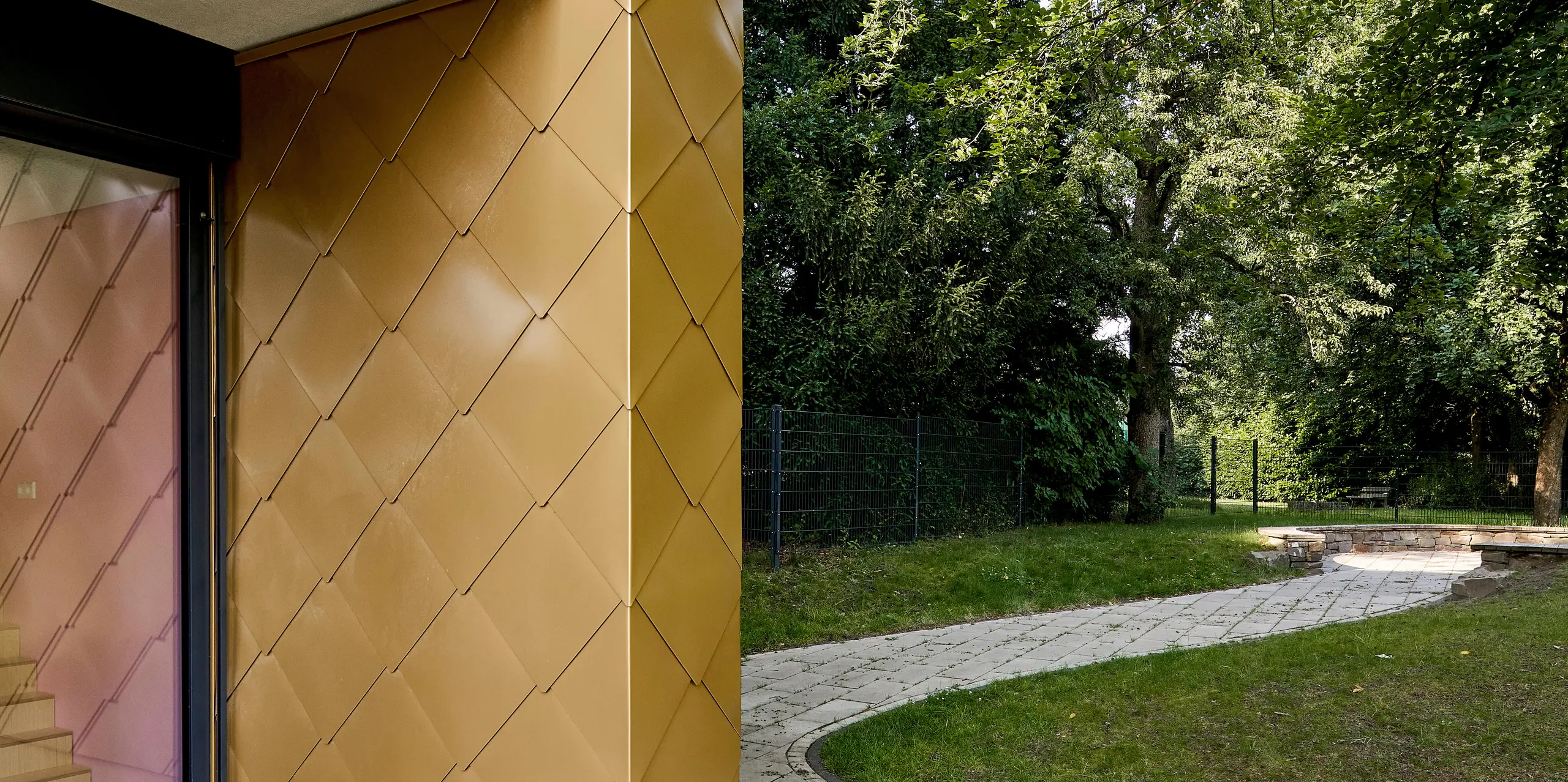 Moderne Architektur mit goldschimmernder PREFA Aluminiumfassade in Schuppenoptik prägt diese dynamische Szene. Die präzise verlegten Rauten reflektieren das Sonnenlicht eindrucksvoll und setzen einen markanten Eyecatcher gegenüber dem grünen Baumbestand. Der elegante Balkon mit klaren Linien ergänzt das hochwertige Erscheinungsbild. Die langlebigen, witterungsbeständigen PREFA Wandrauten überzeugen durch ihre Farbbrillanz, Robustheit und Wartungsfreiheit – ideal für anspruchsvolle, nachhaltige Fassadengestaltung in urbaner und naturnaher Umgebung.