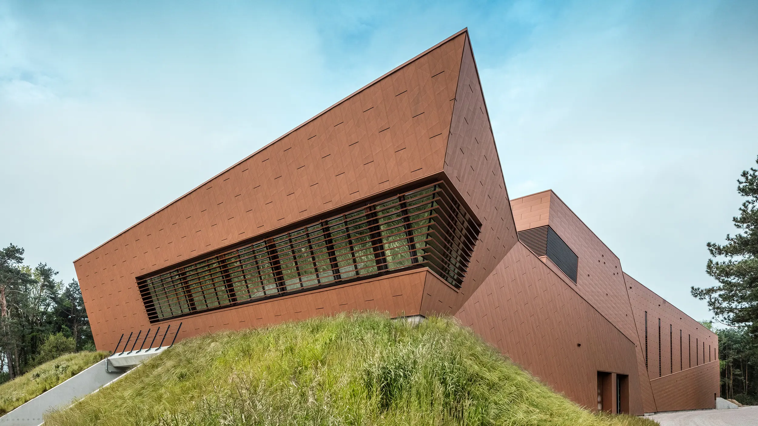 Markantes Trinkwasserpumpwerk von WML in Lottum mit skulptural geneigter Gebäudeform und prägnanter PREFA Siding.X Aluminiumfassade in warmem Rostton. Die dynamisch strukturierte Oberfläche reflektiert Licht und setzt starke architektonische Akzente im Landschaftsraum. Großzügige Lamellenöffnungen und klare Linienführung verbinden Technik und Gestaltung. Die langlebigen, leichten und korrosionsbeständigen PREFA Aluminiumpaneele überzeugen durch nachhaltige Recyclingfähigkeit, hohe und eine wartungsarme Gebäudehülle – ein moderner Eyecatcher für zukunftsorientierten Infrastrukturbau.