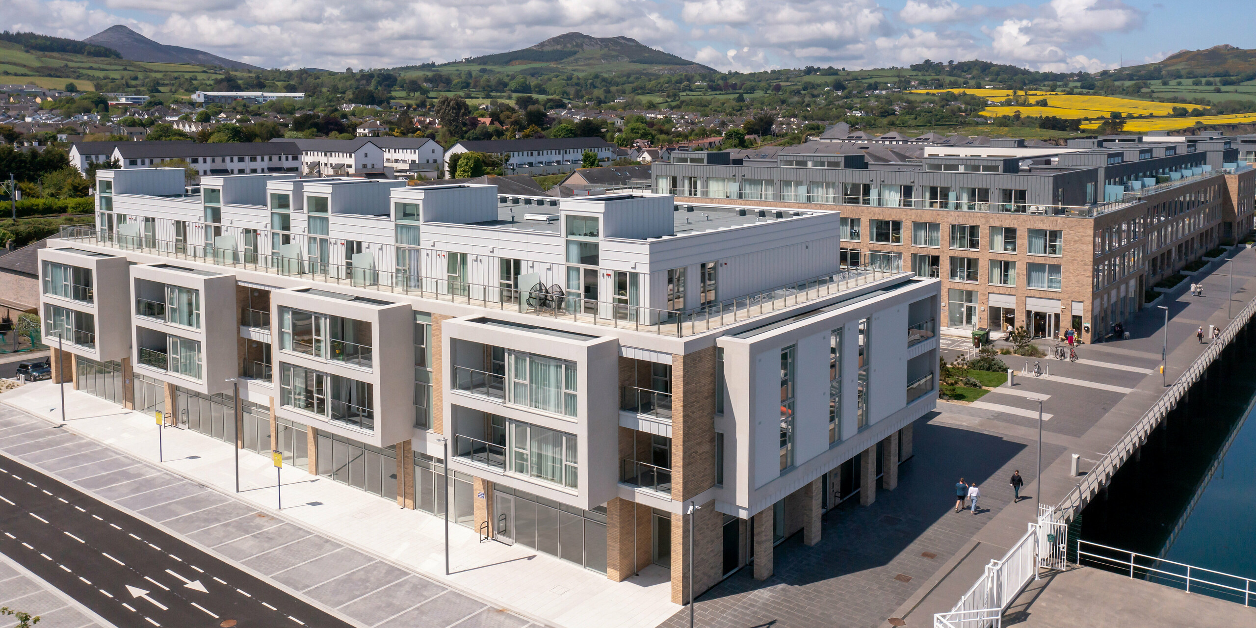 Marina Village Greystones in Wicklow, Ireland - wide angle view of a modern residential development directly on the waterfront with a view of the marina. The building complex is clad with approx. 2,000 m² of robust PREFALZ in the colours P.10 Prefa White and P.10 Light Grey. The elegant architecture includes large glass fronts, spacious balconies and an aesthetic façade design. Surrounded by a picturesque landscape and high-quality sheet metal from PREFA, the development offers lasting protection and timeless aesthetics for exclusive living by the sea.