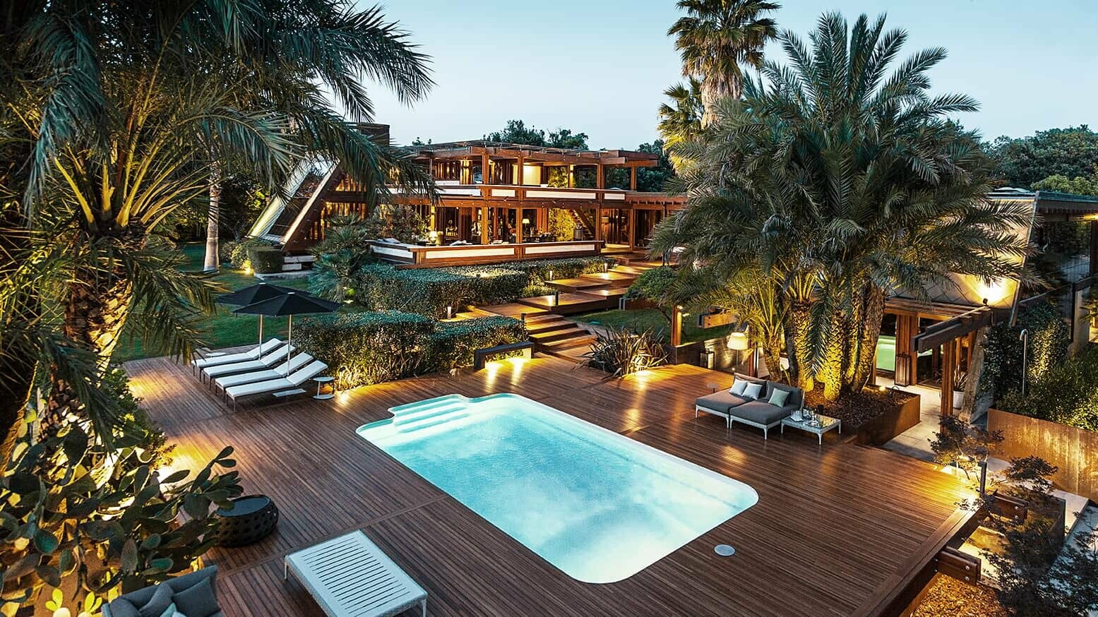 Wide angle view of the Pagano complex with pool and comfortable sunbathing areas. Palm trees and indirect light give the whole a luxurious flair.
