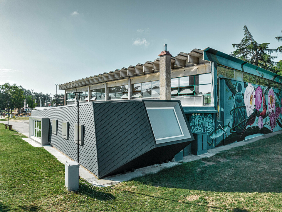 The Rodriguez gym and its new extension with the scaly shomboid skin made of PREFA aluminium from the side, the lawn extending before it and on the side. Part of a sports field can be seen in the background.