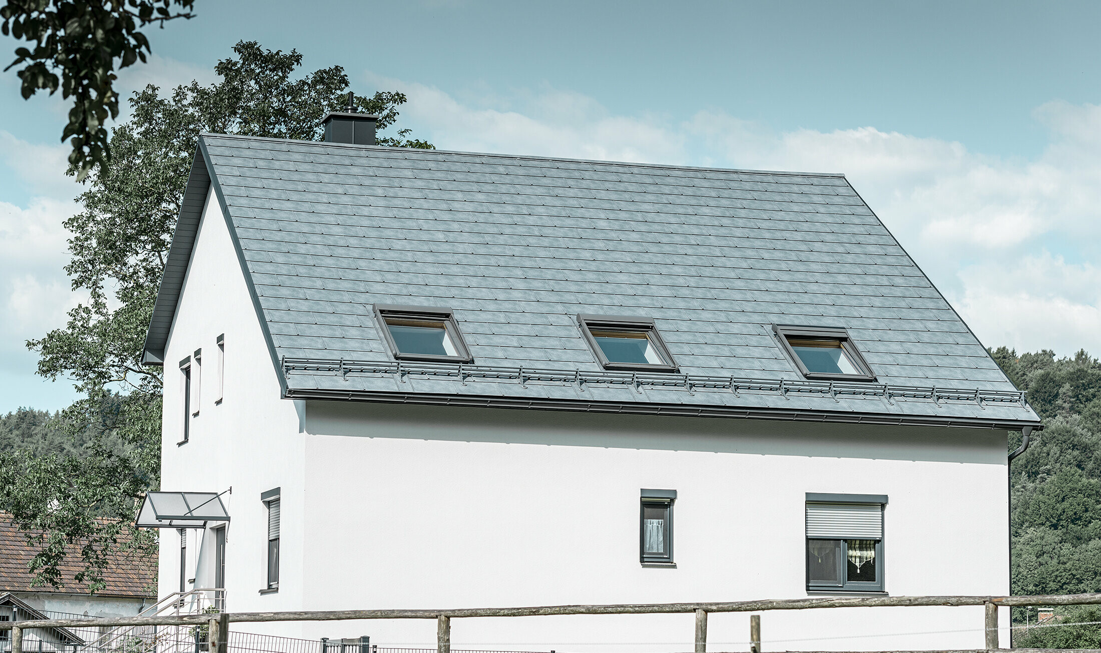 The gabled roof of the classic detached house was covered with the new PREFA R.16 roof tile in stone grey. Three roof windows were inserted in the roof area and snow guards were installed. The façade is kept simple in white.