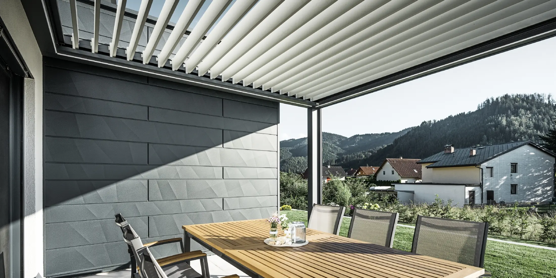 Sheltered terrace covered with slats, with table and chairs; one side of the façade is clad with PREFA Siding.X façade panels in anthracite.