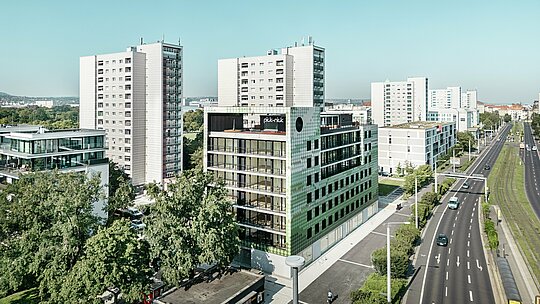 Aerial view of the residential and commercial building ‘Pick-Nick’ at Straßburger Platz in Dresden with the striking PREFA façade made of aluminium rhomboids in various shades of green and beige. The building is located on a main road, embedded in a modern urban environment with high-rise buildings and green spaces. The striking architecture and the high-quality PREFA façade set visual accents in the cityscape.