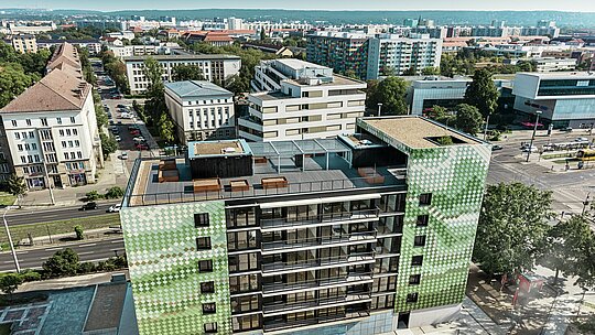 Aerial view of the ‘Pick-Nick’ residential and commercial building in Dresden with a PREFA façade made of rhomboid tiles in various shades of green and beige. The impressive city panorama can be seen in the background. The building has a spacious roof terrace with flowerbeds and a children's playground. Embedded in an urban environment, it offers a combination of modern architecture, sustainability and functional design.