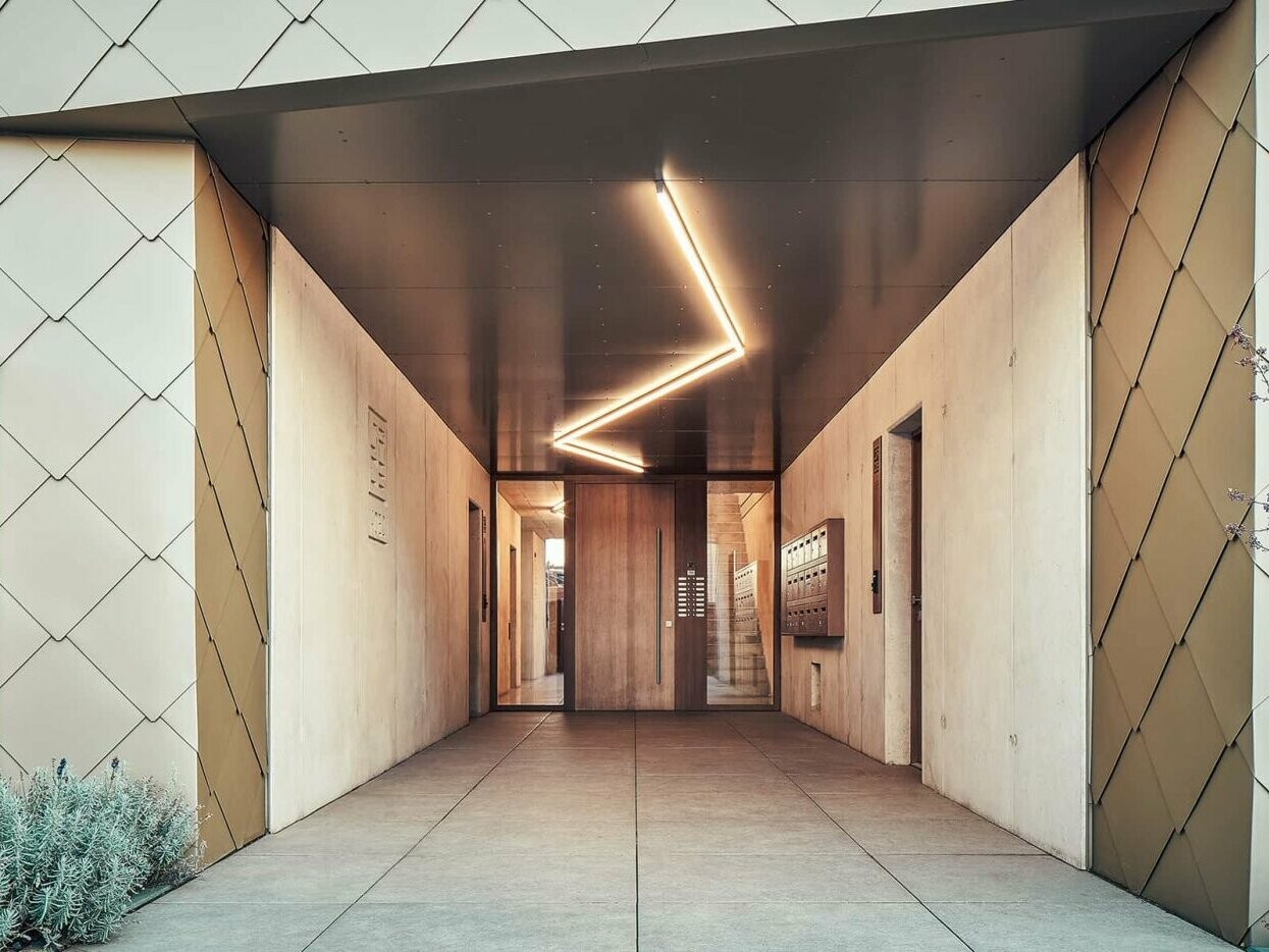 The picture shows the strikingly illuminated entrance to the apartment building with concrete and façade elements from PREFA.