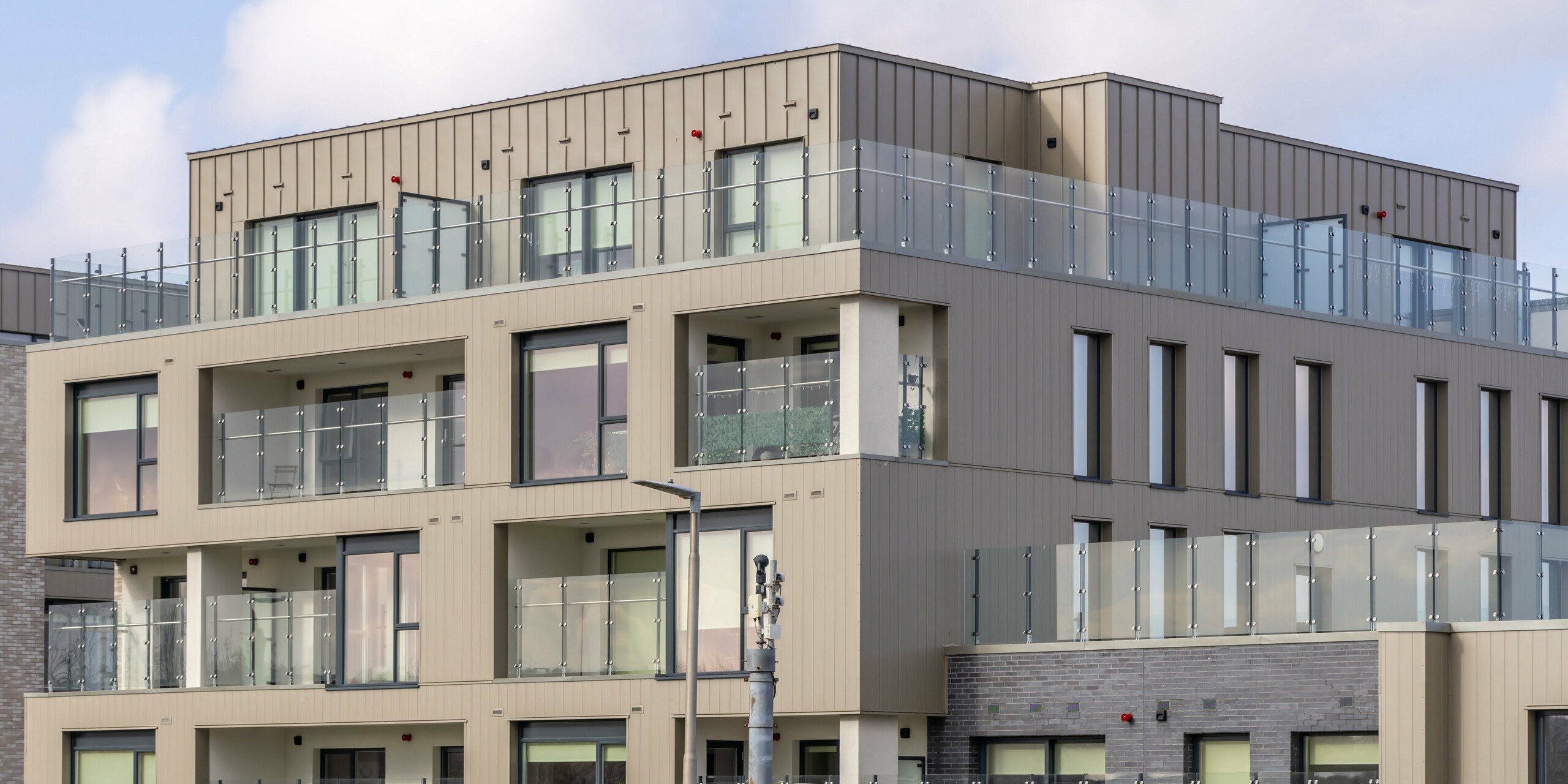 Palmers Gate residential complex in Palmerstown, Dublin, Ireland - close-up view of a residential building with bronze-coloured PREFA sidings and PREFALZ façade system. The balconies are equipped with elegant glass balustrades, which emphasise the modern living comfort. The façade combines bronze-coloured aluminium cladding with brick elements, creating a harmonious and attractive appearance. The building complex provides residents with high quality outdoor spaces and boasts a robust and aesthetically pleasing architecture.