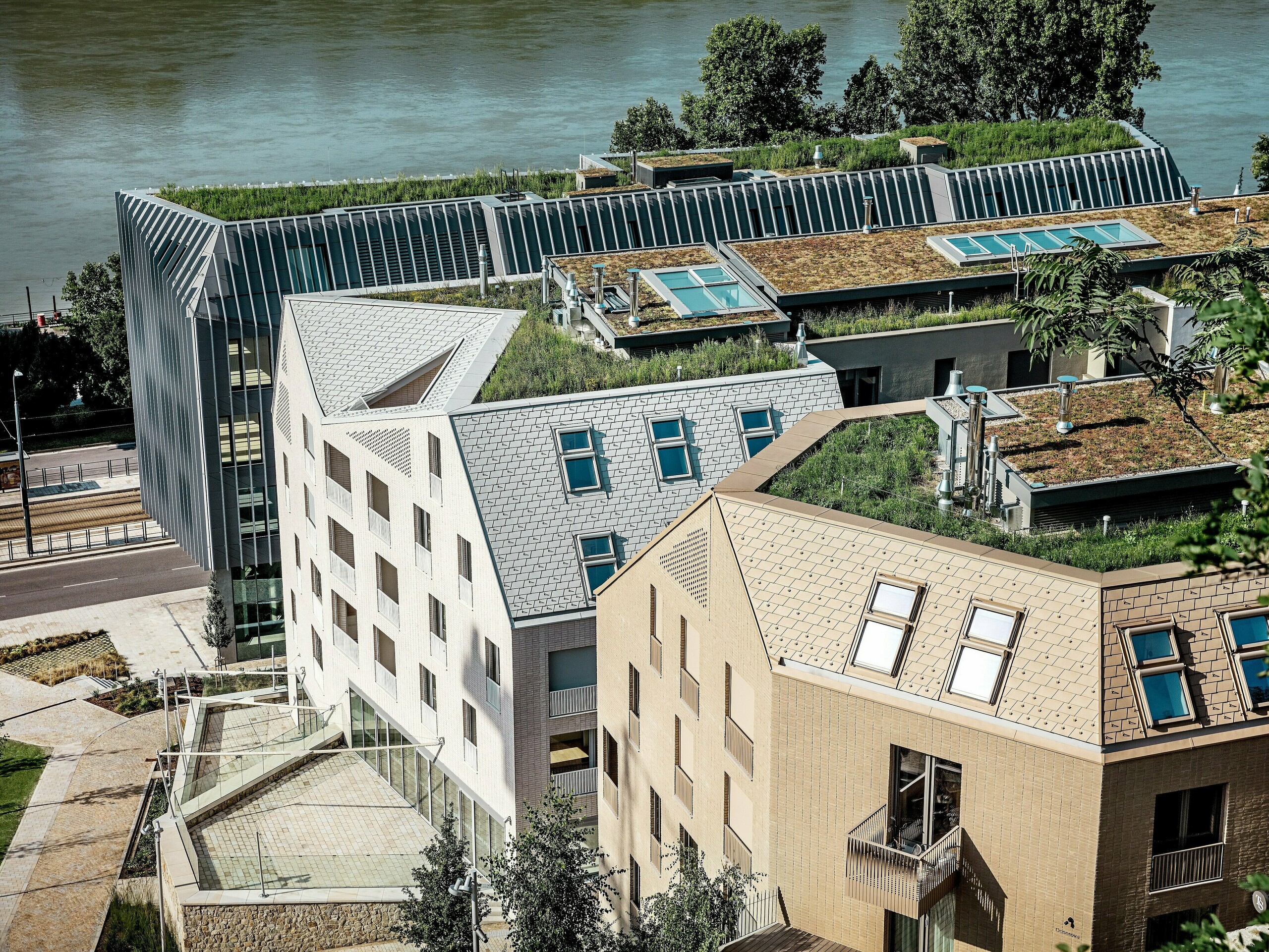 Modernes Stadtquartier in Bratislava mit markanter Architektur und begrünten Dachlandschaften direkt am Flussufer: Die filigranen, steil geneigten Dächer und Gauben werden von hochwertigen PREFA Aluminium Dachschindeln in Prefaweiß, Sandbraun und Bronze geprägt und ziehen sofort alle Blicke auf sich. Großzügige Dachflächenfenster, klare Linien und üppige Dachbegrünung verbinden Design und Nachhaltigkeit. Die langlebigen, wartungsarmen PREFA Aluminiumlösungen überzeugen durch Widerstandskraft, Leichtigkeit und Witterungsbeständigkeit – ideal für anspruchsvolle urbane Bauprojekte.