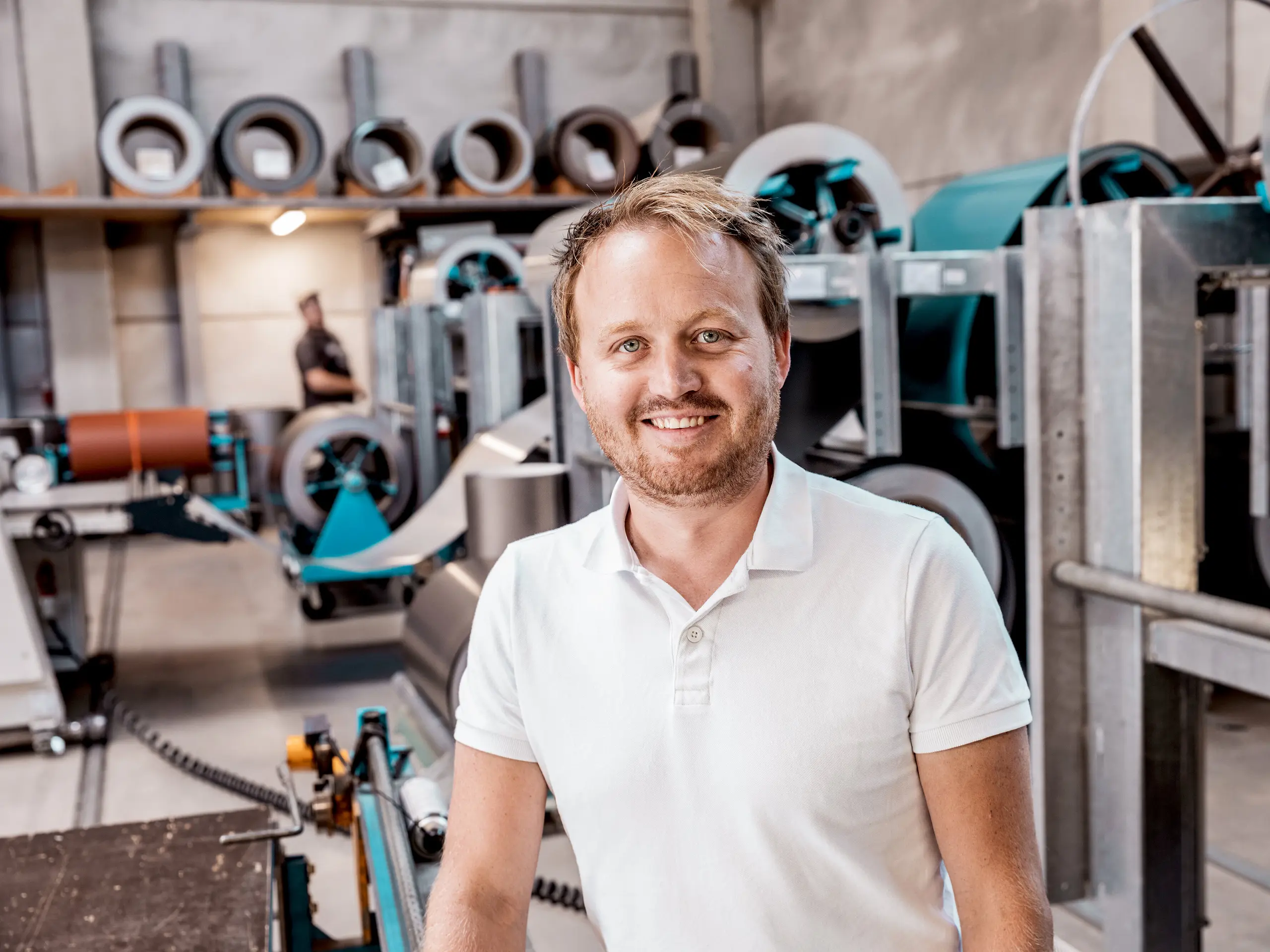 Das Bild zeigt David Thumfart, Spengler bei der Spengler- und Dachdeckerbetriebes Kapl Bau GmbH, in seiner Werkstatt vor den Maschinen zur Verarbeitung von Metallrollen. Mit einem Lächeln und einem professionellen Auftreten steht er stolz inmitten der hochmodernen Produktionsausstattung, die für die Fertigung von Fassaden- und Dachsystemen aus Aluminium genutzt wird. Die Maschinen im Hintergrund verdeutlichen die Präzision, die für die Herstellung hochwertiger Produkte wie die PREFALZ-Fassade erforderlich ist. Diese wurde im Einfamilienhaus in Freistadt verwendet. Thumfart verkörpert damit das Engagement und die Expertise, die in die Herstellung und Montage langlebiger und ästhetischer Baukomponenten fließen.