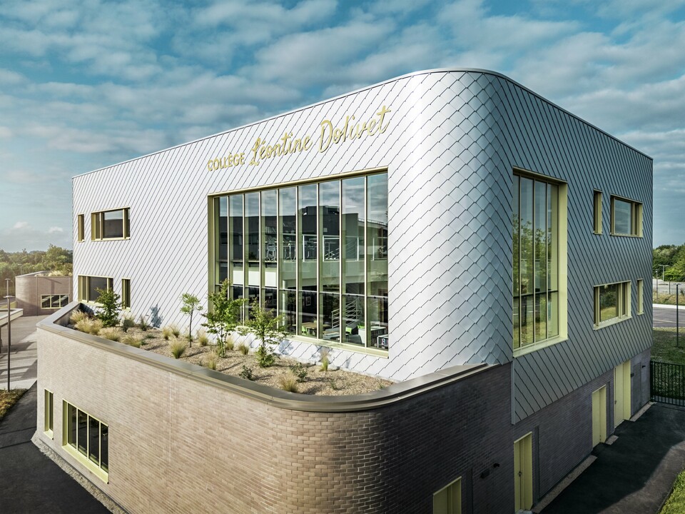 Das Foto zeigt eine markante Gebäudeecke des Collège Léontine Dolivet in Cesson-Sévigné. Die Fassade besteht aus silbermetallic PREFA Wandrauten, die in einem diagonalen Muster angeordnet sind und das Licht reflektieren. Auffällig ist die geschwungene Form der oberen Etage, die dem Bau eine weiche, moderne Anmutung verleiht. Große Glasfronten öffnen den Blick ins Innere, während gelbe Fensterrahmen lebendige Akzente setzen. Im Vordergrund befindet sich ein bepflanzter Bereich mit Kies und Sträuchern, der die Architektur harmonisch ergänzt. Der Himmel ist leicht bewölkt, was die metallische Oberfläche betont.