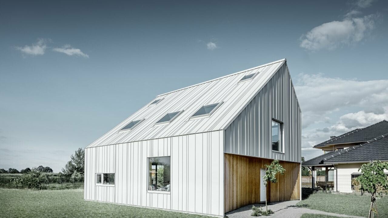 Modern detached house with gable roof, the entire outer skin is clad with PREFALZ in PREFA white. The panels of different widths extend from the roof over the façade. Several roof windows are installed in the roof.