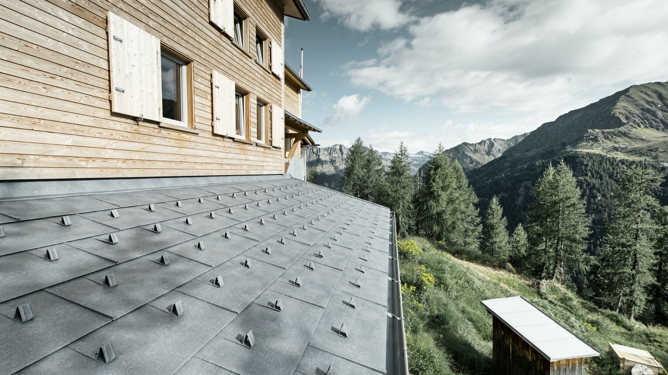 Close-up of a canopy on a wooden cabin. The canopy is covered with PREFA FX.12 aluminium panels in stone grey with snow guards. The gutter is held in place by overbars.