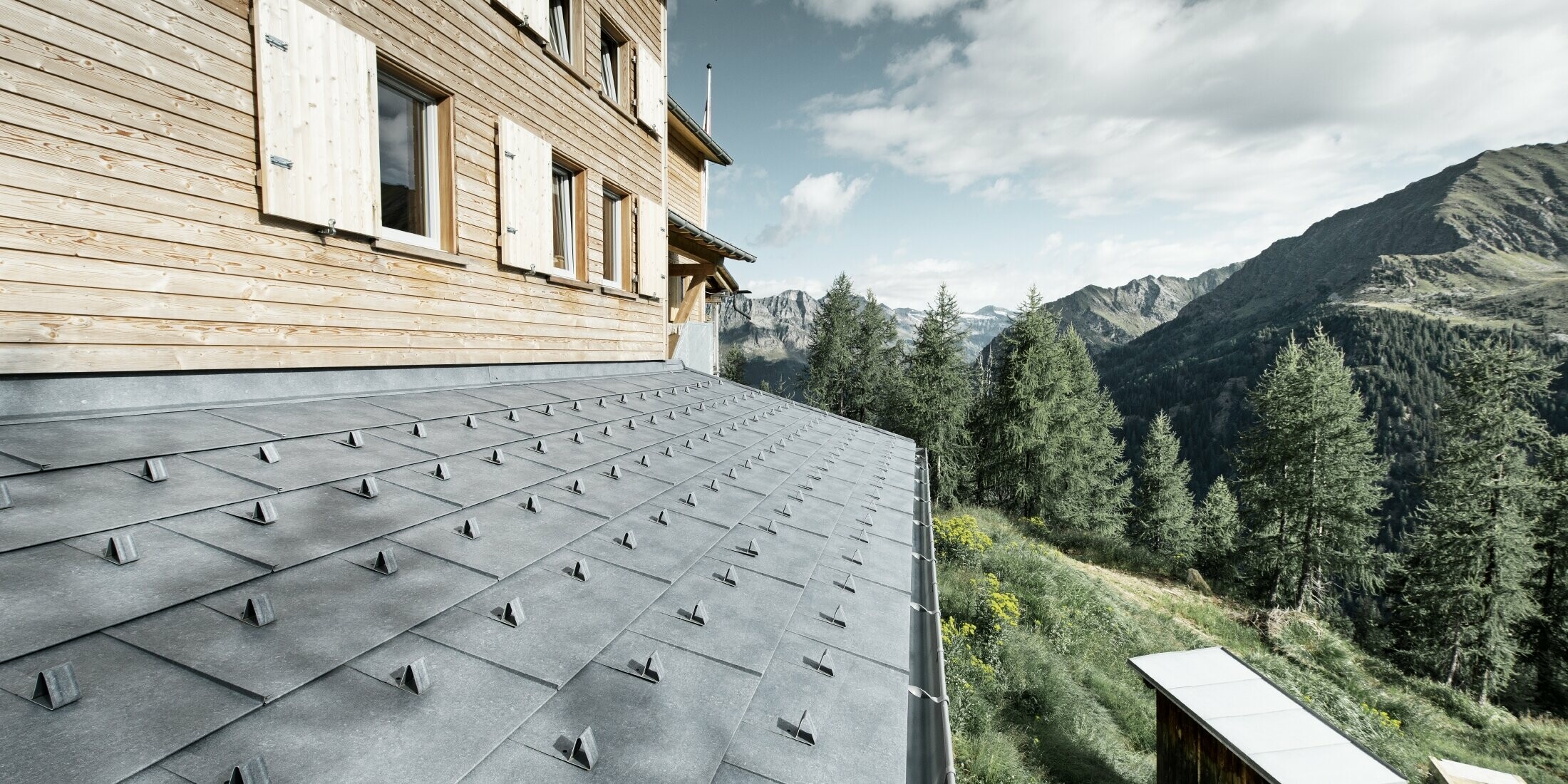 Close-up of a canopy on a wooden cabin. The canopy is covered with PREFA FX.12 aluminium panels in stone grey with snow guards. The gutter is held in place by overbars.