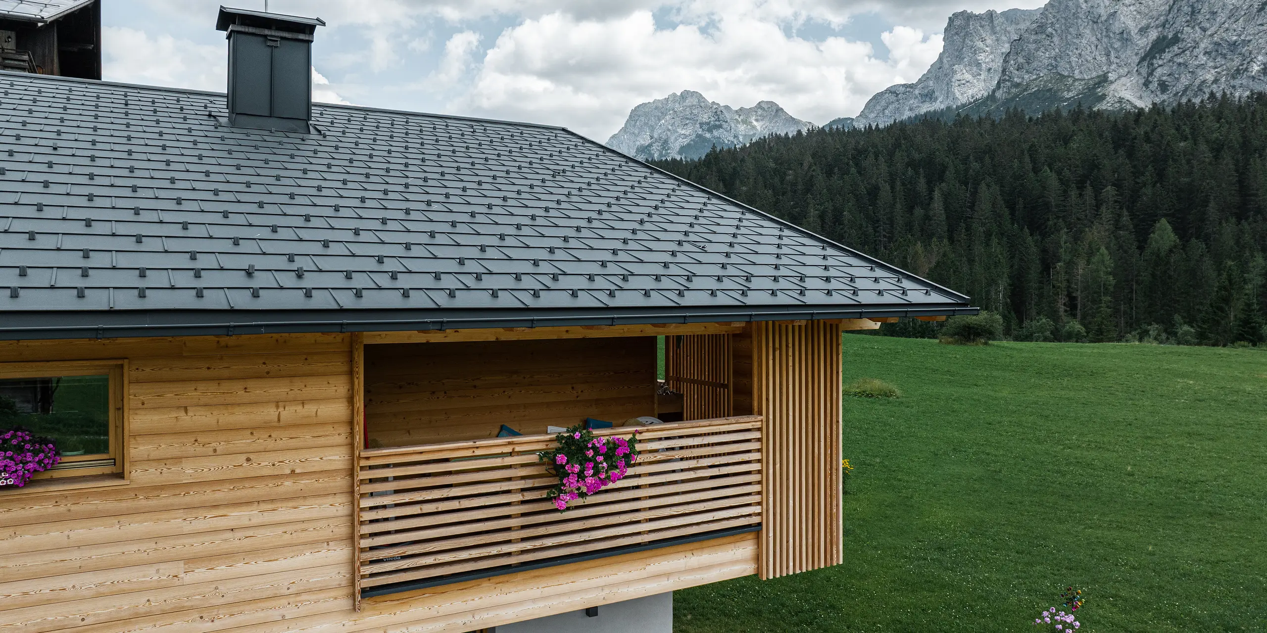 Die malerische Seitenansicht der Loggia von Casa Hoffe in Sappada, Italien, zeigt die harmonische Integration von PREFA Dachplatten R.16 in P.10 Schwarz mit einer naturbelassenen Holzfassade. Die traditionelle Bauweise der Loggia, kombiniert mit der modernen, langlebigen Dachlösung, fügt sich nahtlos in die atemberaubende Bergkulisse der Dolomiten ein und unterstreicht das naturnahe Wohnkonzept der Region.