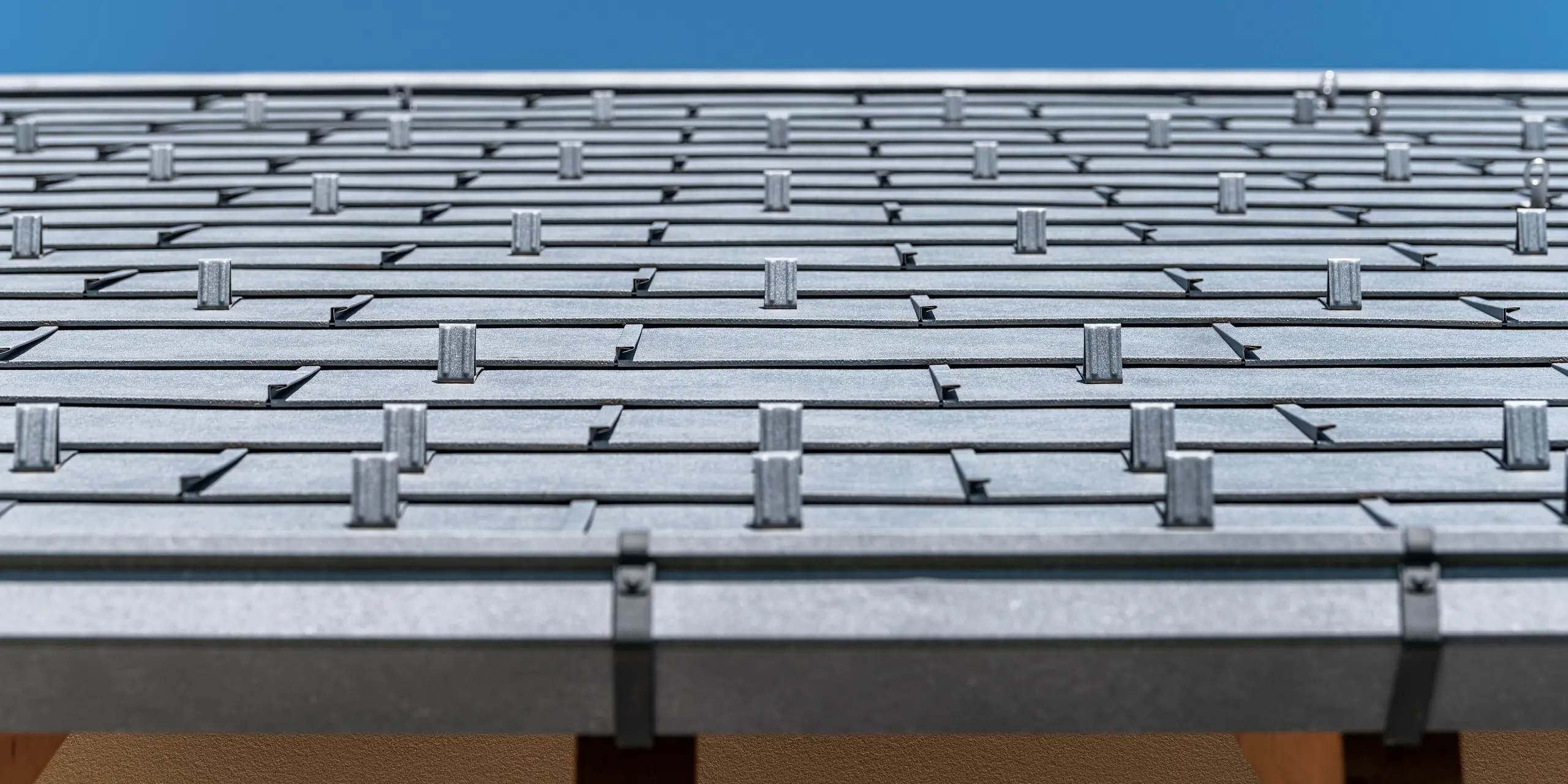 Detailansicht des PREFA Daches eingedeckt mit Dachpaneel R.16 in Farbe P.10 anthrazit. Das Bild zeigt präzise verarbeitete Aluminium-Dachplatten. Die Aufnahme hebt die markante Struktur und das moderne Design des Daches hervor, das im Kontrast zum strahlend blauen Himmel steht und die innovative und langlebige Dachlösung von PREFA unterstreicht.
