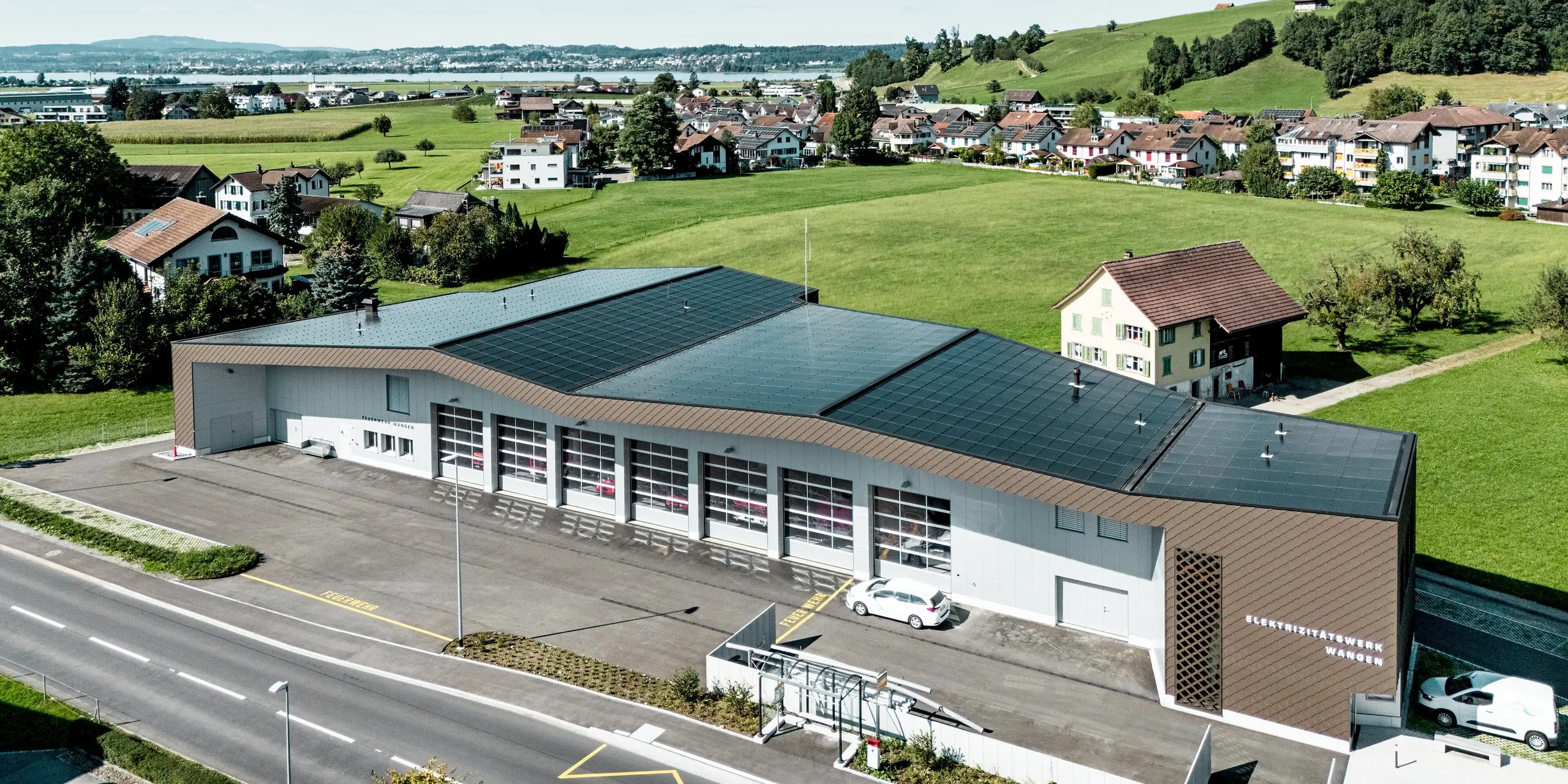 Luftaufnahme des Infrastrukturgebäudes für die Feuerwehr und des Elektrizitätswerks in Wangen in der Schweiz, eingebettet in eine malerische Hügellandschaft. Die markante Fassadenbekleidung aus PREFA Wandrauten 29 × 29 in der Farbe P.10 Braun setzt architektonische Akzente und harmoniert mit den hellen Aluminium-Verbundplatten in der Farbe P.10 Prefaweiß. Die langlebigen Aluminiumprodukte überzeugen durch ihre hohe Witterungsbeständigkeit, ihren geringen Wartungsaufwand und ihre moderne, nachhaltige Gebäudehülle.