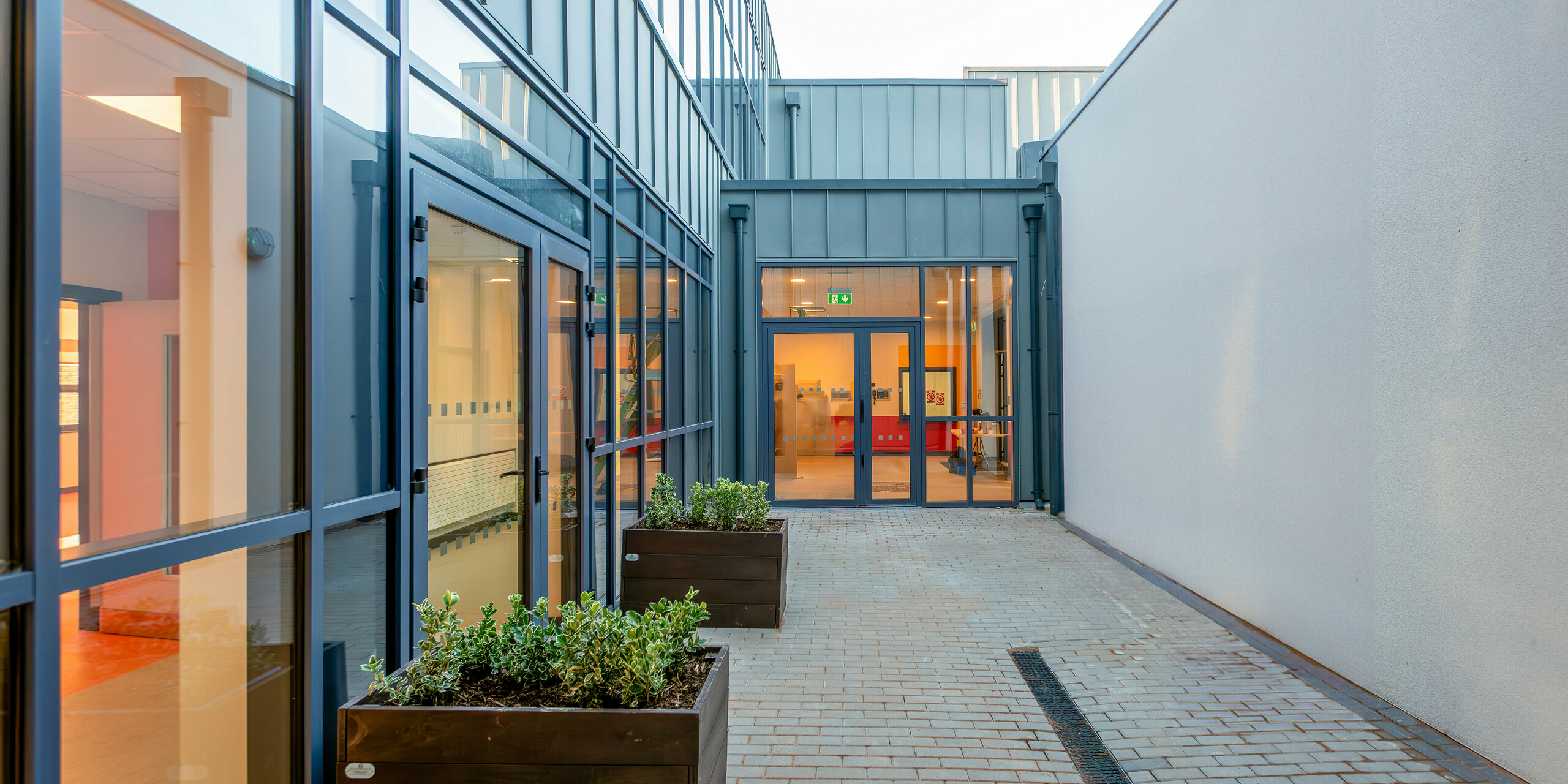 Modern passageway of Scoil Mhuire Naofa in Carrigtwohill, Republic of Ireland. Large glass fronts and a PREFALZ aluminium facade in P.10 light grey ensure a light-flooded, contemporary architecture. Planters and light-coloured materials create a welcoming atmosphere.