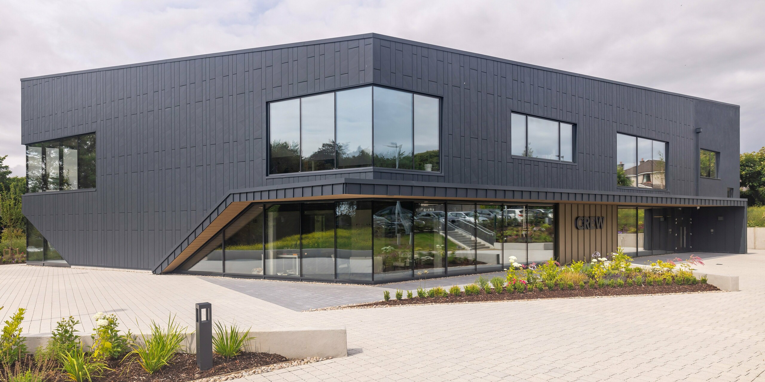 Front view of the CREW building in Galway, Ireland, with modern PREFA aluminium façade made of Siding.X and PREFALZ in P.10 anthracite. The angled lines and large window areas characterise the architecturally sophisticated design of the building. The anthracite-coloured aluminium cladding in combination gives the building an elegant and contemporary aesthetic, which is complemented by the neat landscaping.