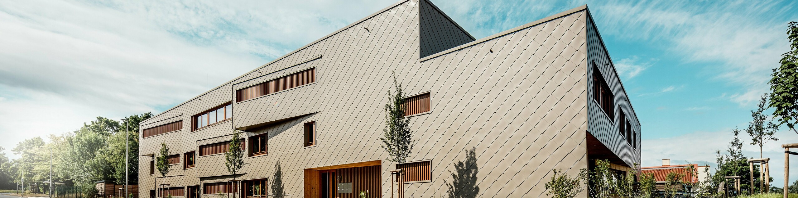 Aus einer markanten Untersichtperspektive präsentiert sich die Kindertagesstätte in Alzenau mit ihrer prägnanten, dynamischen Linienführung und der hochwertigen PREFA Aluminiumfassade in Bronze. Die diagonale Anordnung der Wandrauten 44 × 44 verleiht dem Baukörper eine klare Struktur und eine elegante, homogene Oberfläche. Versetzte Fensteröffnungen und vertikale Holzlamellen setzen rhythmische Akzente, während die reduzierte Formensprache moderne Zurückhaltung signalisiert. Die Kombination aus langlebigem Aluminium, natürlichem Holz und einem durchdachten Grünkonzept entlang der Straße unterstreicht den hohen Anspruch an Nachhaltigkeit und Bauqualität. Dieses Bild zeigt eindrucksvoll, wie funktionale Architektur mit PREFA Produkten gestalterisch überzeugen und städtebaulich Maßstäbe setzen kann.