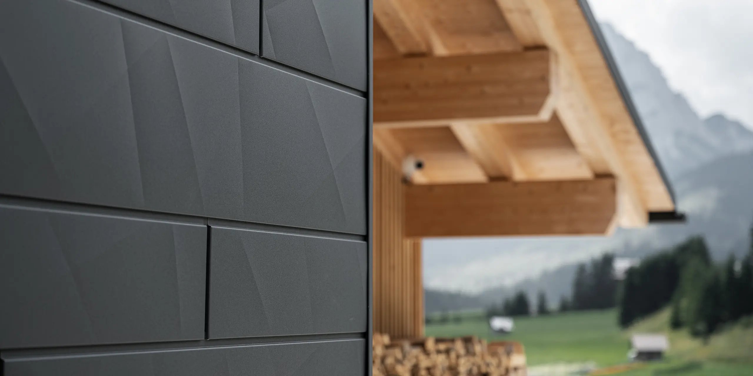 Close-up of PREFA Siding.X façade panels in P.10 black on Casa Hoffe in Sappada, Italy. The aluminium façade system is skillfully installed in a modern, abstract design, creating an appealing contrast with the visible wooden elements of the roof structure. The innovative façade solution blends seamlessly into the alpine setting and represents a combination of functionality, durability and aesthetic building culture.