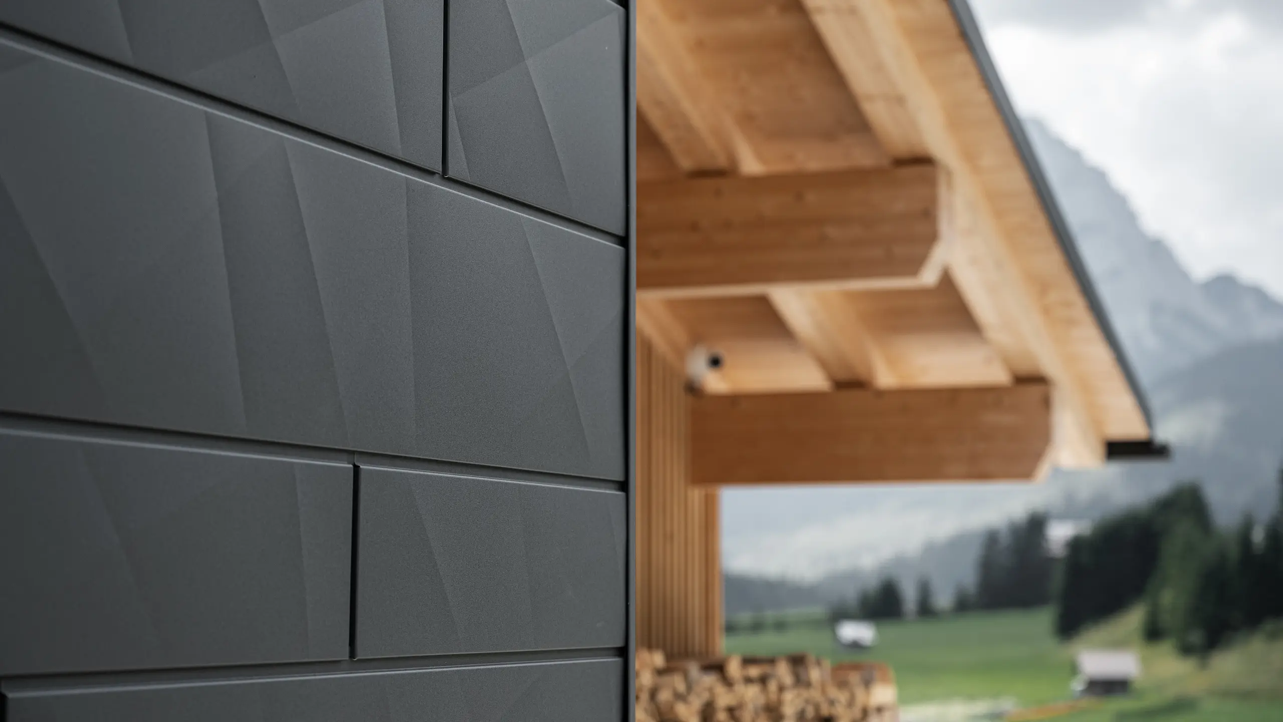 Close-up of PREFA Siding.X façade panels in P.10 black on Casa Hoffe in Sappada, Italy. The aluminium façade system is skillfully installed in a modern, abstract design, creating an appealing contrast with the visible wooden elements of the roof structure. The innovative façade solution blends seamlessly into the alpine setting and represents a combination of functionality, durability and aesthetic building culture.
