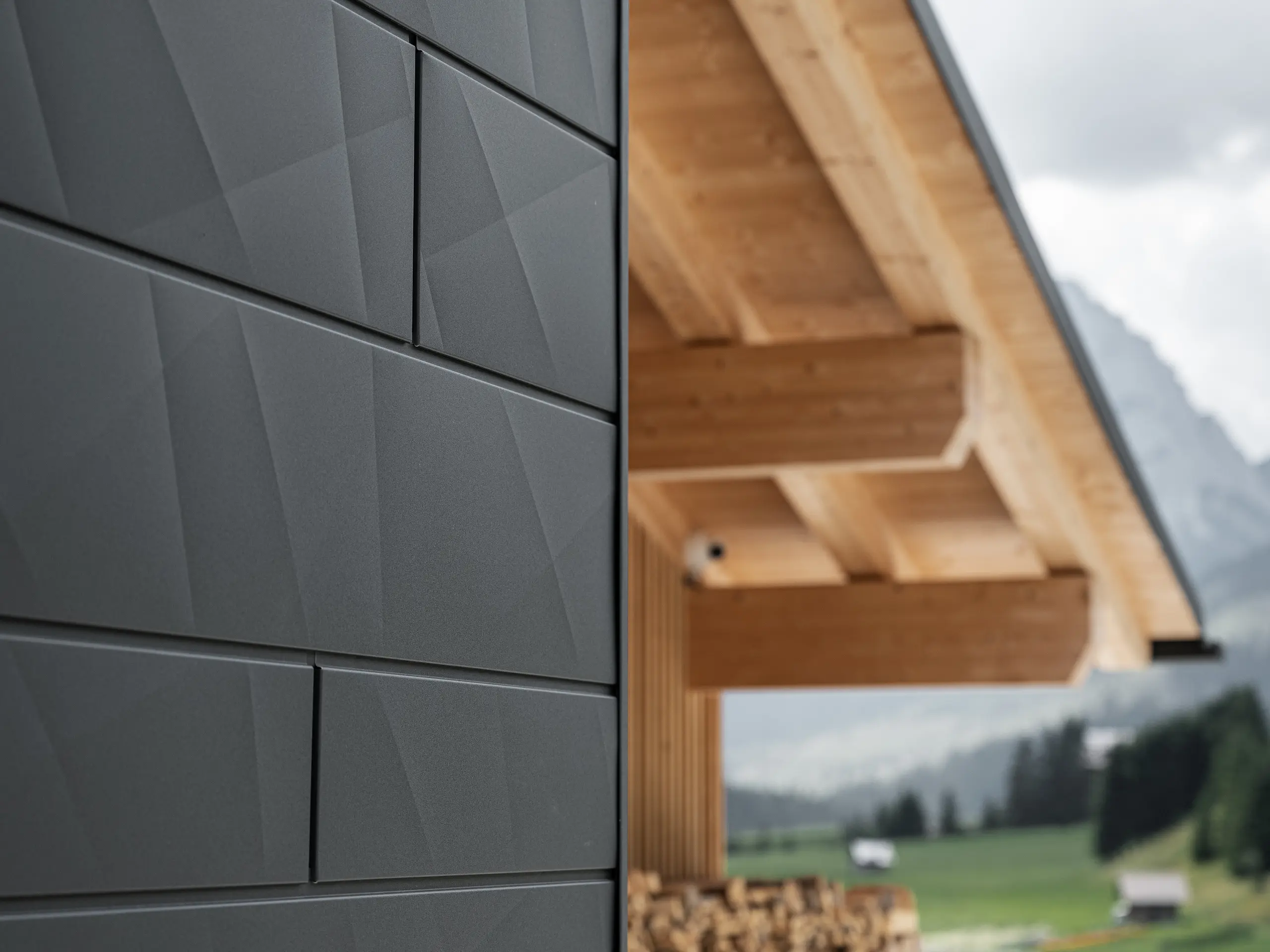 Close-up of PREFA Siding.X façade panels in P.10 black on Casa Hoffe in Sappada, Italy. The aluminium façade system is skillfully installed in a modern, abstract design, creating an appealing contrast with the visible wooden elements of the roof structure. The innovative façade solution blends seamlessly into the alpine setting and represents a combination of functionality, durability and aesthetic building culture.