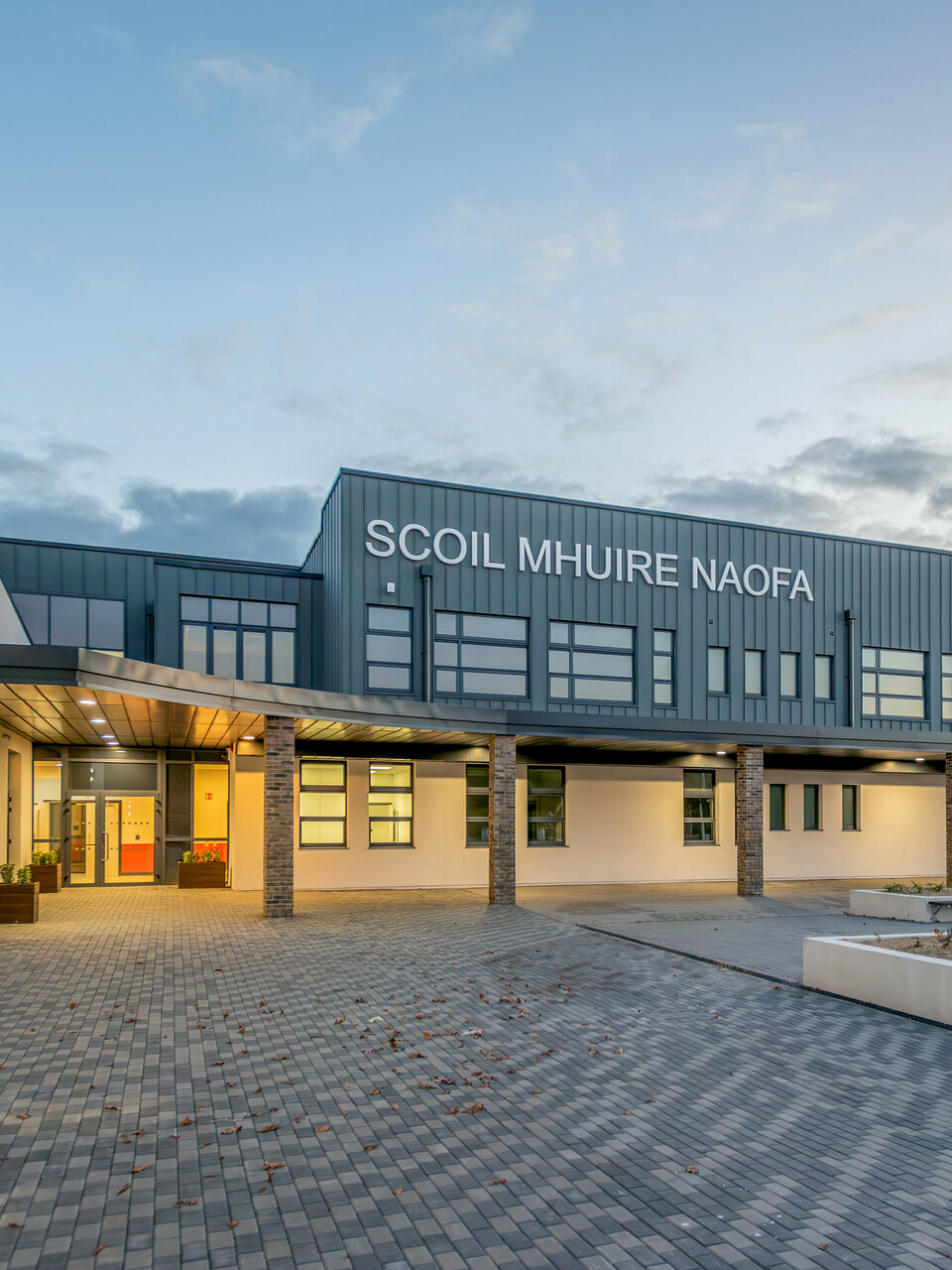 The Scoil Mhuire Naofa in Carrigtwohill, Ireland, at dusk. The contemporary façade made of PREFALZ aluminium in the colour P.10 light grey, combined with brick columns and clean lines, gives the school building an inviting and high-quality appearance. The clear architecture, stylish lighting and well-kept forecourt create a pleasant atmosphere.