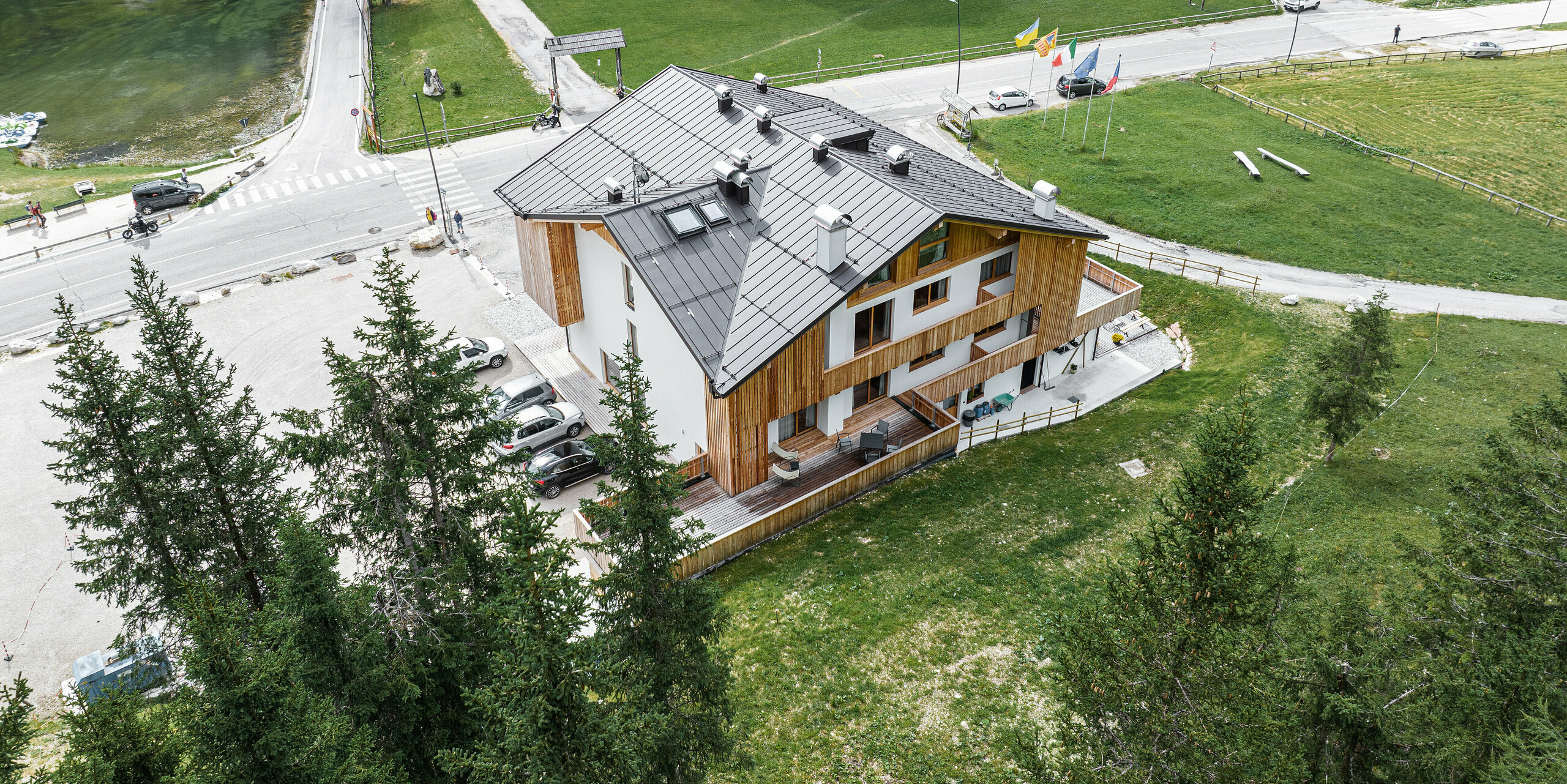 Luftaufnahme des Chalets Alpenrose in Auronzo di Cadore, BL (Italien). Das Gebäude liegt inmitten einer malerischen Berglandschaft, ausgestattet mit einem PREFALZ Dach in der Farbe P.10 Nussbraun. Das moderne Chalet kombiniert traditionelle Holzarchitektur mit einem robusten und langlebigen PREFA Dachsystem, das ideal auf die natürliche Umgebung abgestimmt ist und neben einem Hotel und einem kleinen See liegt. 