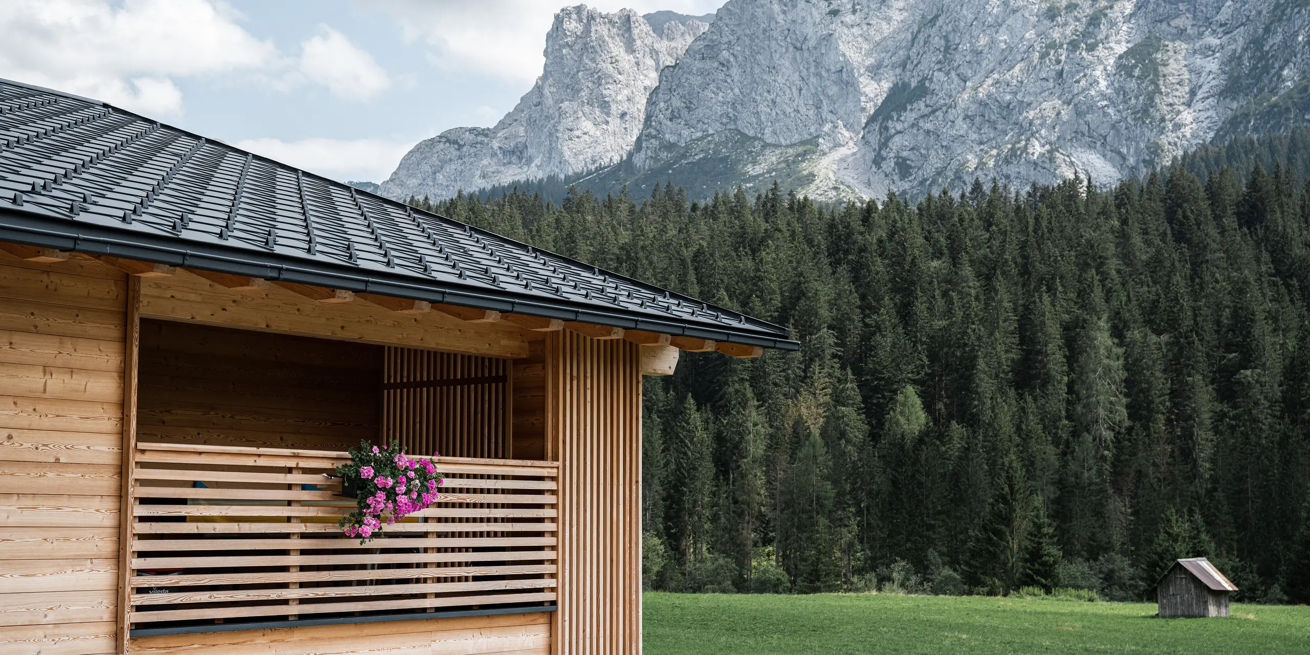 Casa Hoffe in Sappada, Norditalien, eingebettet in die Dolomiten, mit einem PREFA Dach aus Dachplatten R.16 in P.10 Schwarz. Die klaren Linien des Daches kontrastieren mit der natürlichen Holzfassade und der Loggia, die mit blühenden Pflanzen geschmückt ist. Dieses Bild vereint moderne Bautechnik mit traditioneller alpiner Wohnkultur, umgeben von beeindruckender Natur.
