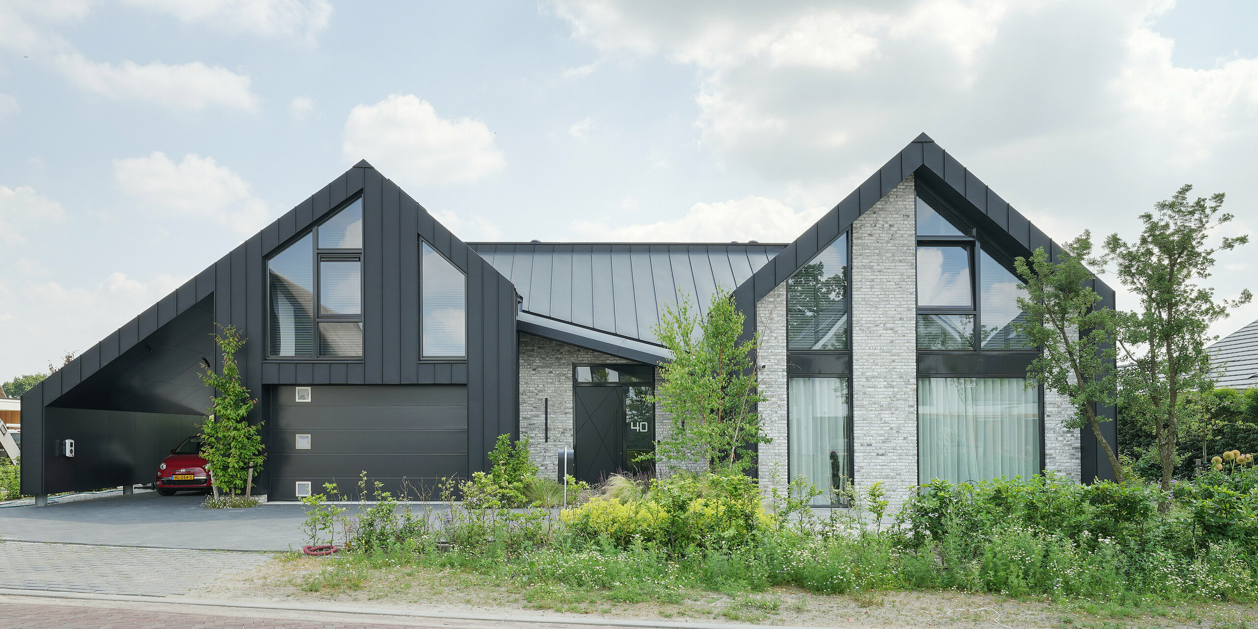 Moderne Wohnarchitektur in Veenendaal, Niederlande, mit dem einzigartigen Zusammenspiel von PREFALZ und einer Fassade, die neben Stehfalzpaneelen aus Aluminium auch Naturstein und große Glasflächen integriert. Das Haus hat markante, spitze Giebel, die dem Entwurf eine dynamische Silhouette verleihen. Das Grundstück ist von einer gepflegten Grünanlage und einer angenehmen Zufahrt umgeben und fügt sich harmonisch in die ruhige Umgebung ein.