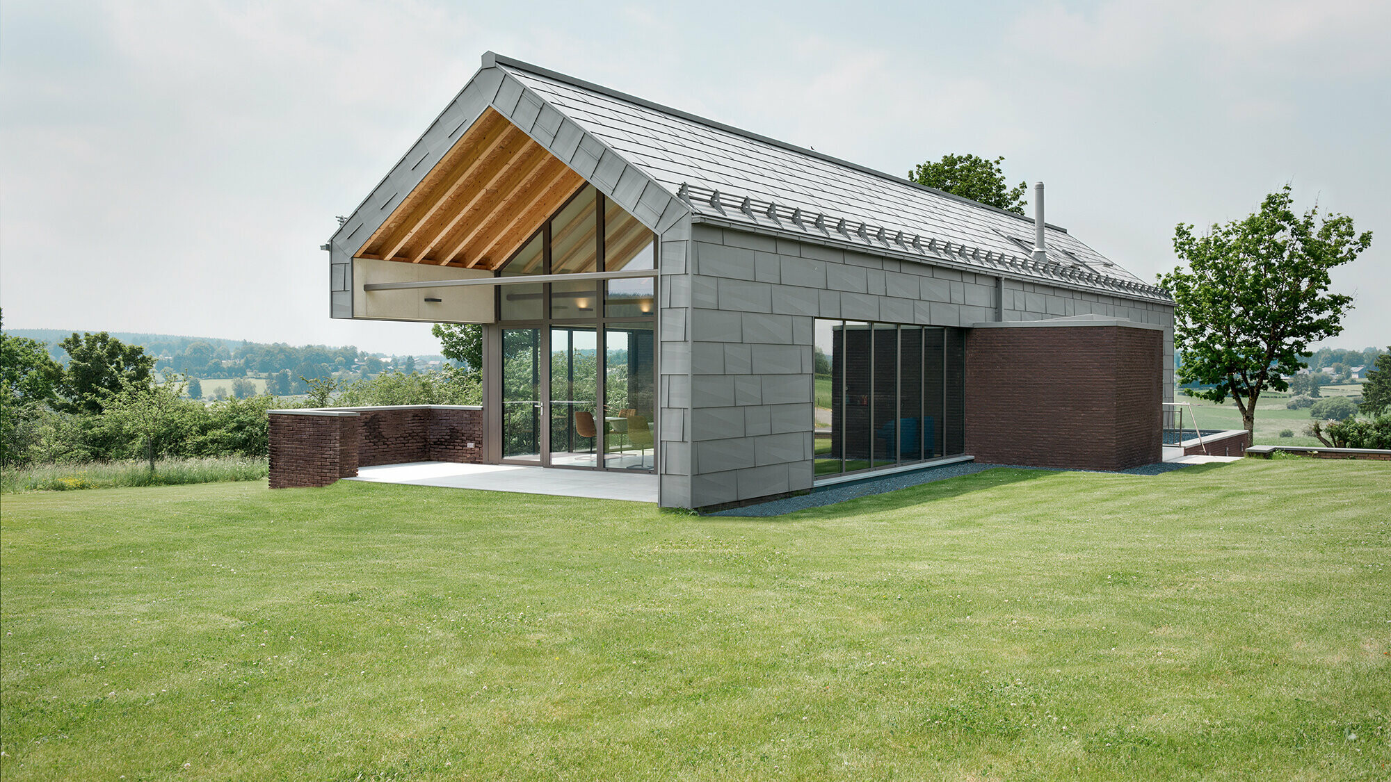 View of the bed and breakfast from the eastern garden side. The glazing is visible, the light grey aluminium envelope is folded around the structure and is framed by brick elements in places.