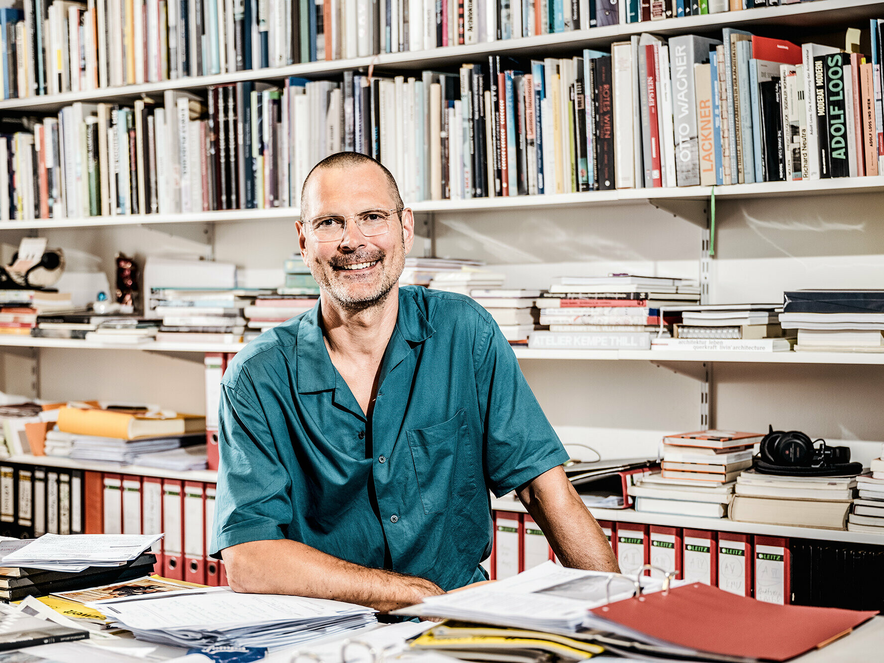 Portrait of architect Eckehart Loidolt in his office room, surrounded by work material and books on a shelf in the background.