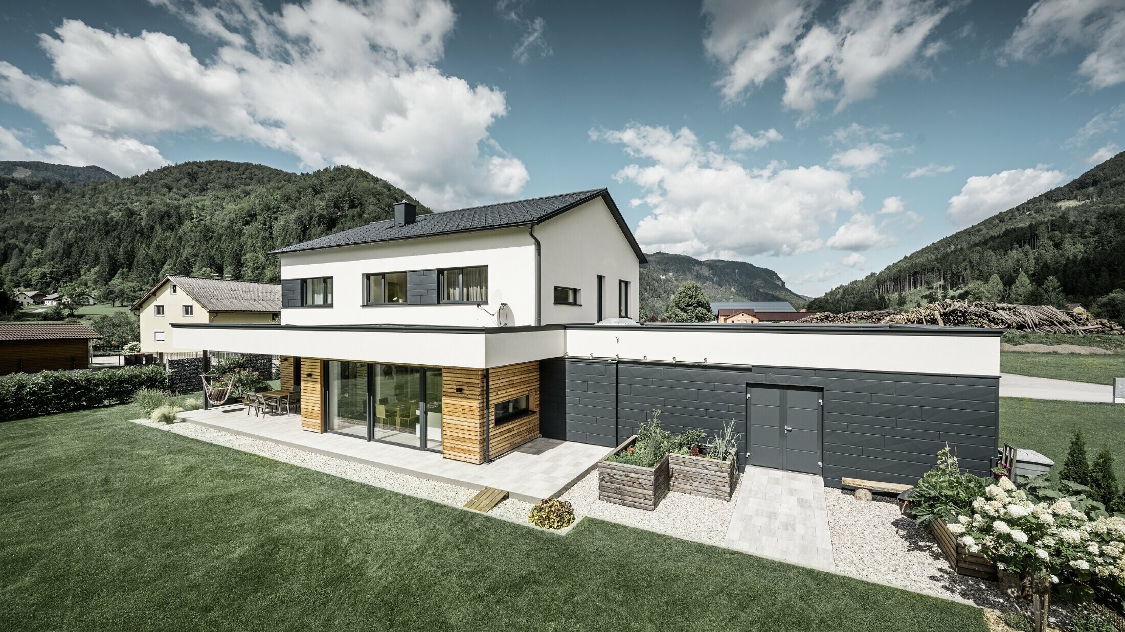 Modern single-family house with white rendered façade and PREFA Siding.X in P.10 anthracite, which forms a striking contrast to the natural environment. Covered with PREFA roof tiles, roof drainage with PREFA half-round gutter and downpipe.