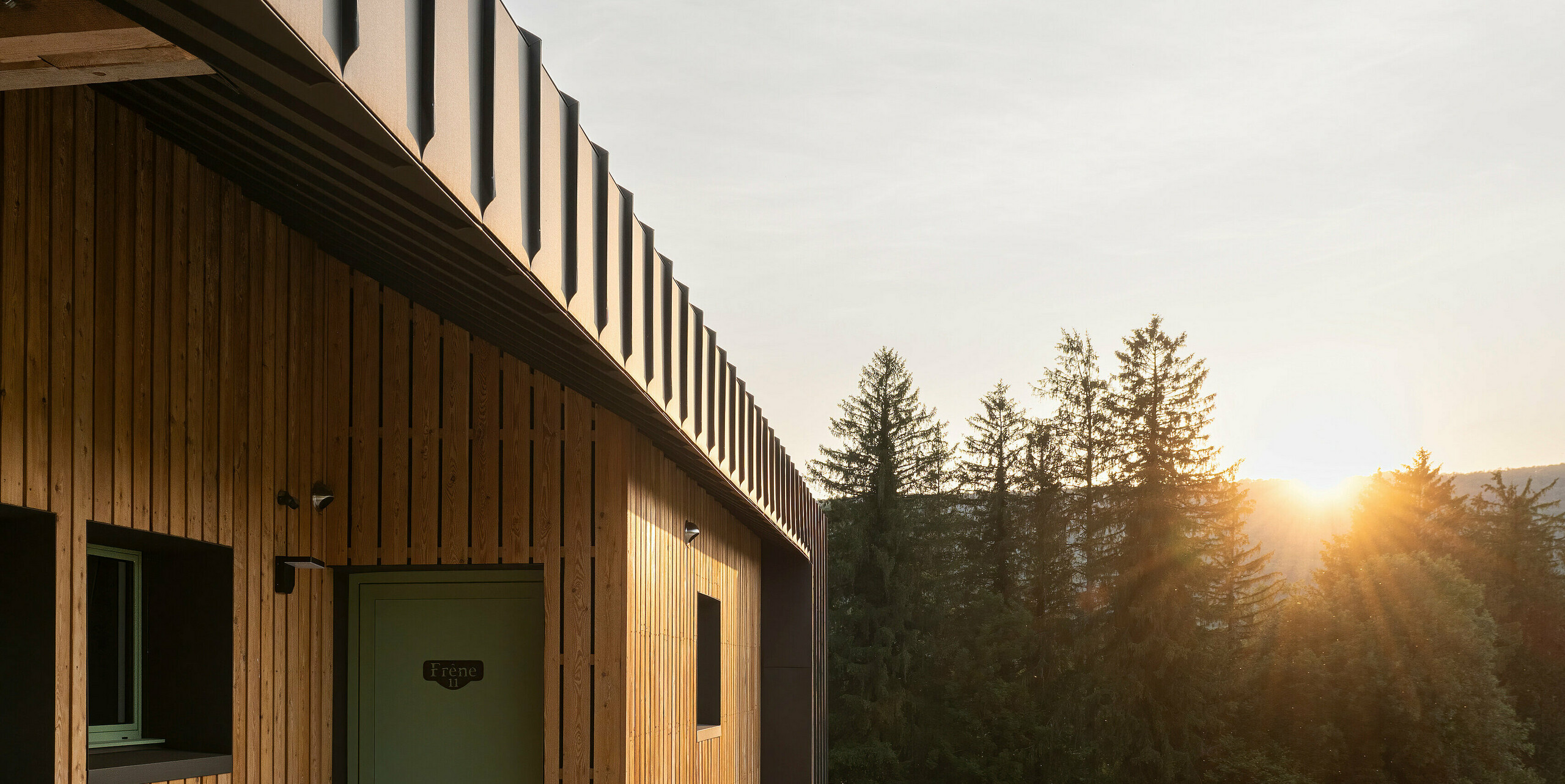 Seitenansicht eines modernen Gebäudes mit Holzfassade und Aluminium-Elementen in der Dämmerung. Die Aluminiumteile am Haus bestehen aus PREFALZ in der Farbe P.10 Braun. Eine Reihe von Stühlen ist vor dem Gebäude neben der Wand aufgestellt. Im Stockwerk darüber verläuft ein Geländer aus Metall. Die Sonne geht hinter den Bäumen am Horizont unter und taucht die Szene in warmes Licht. 