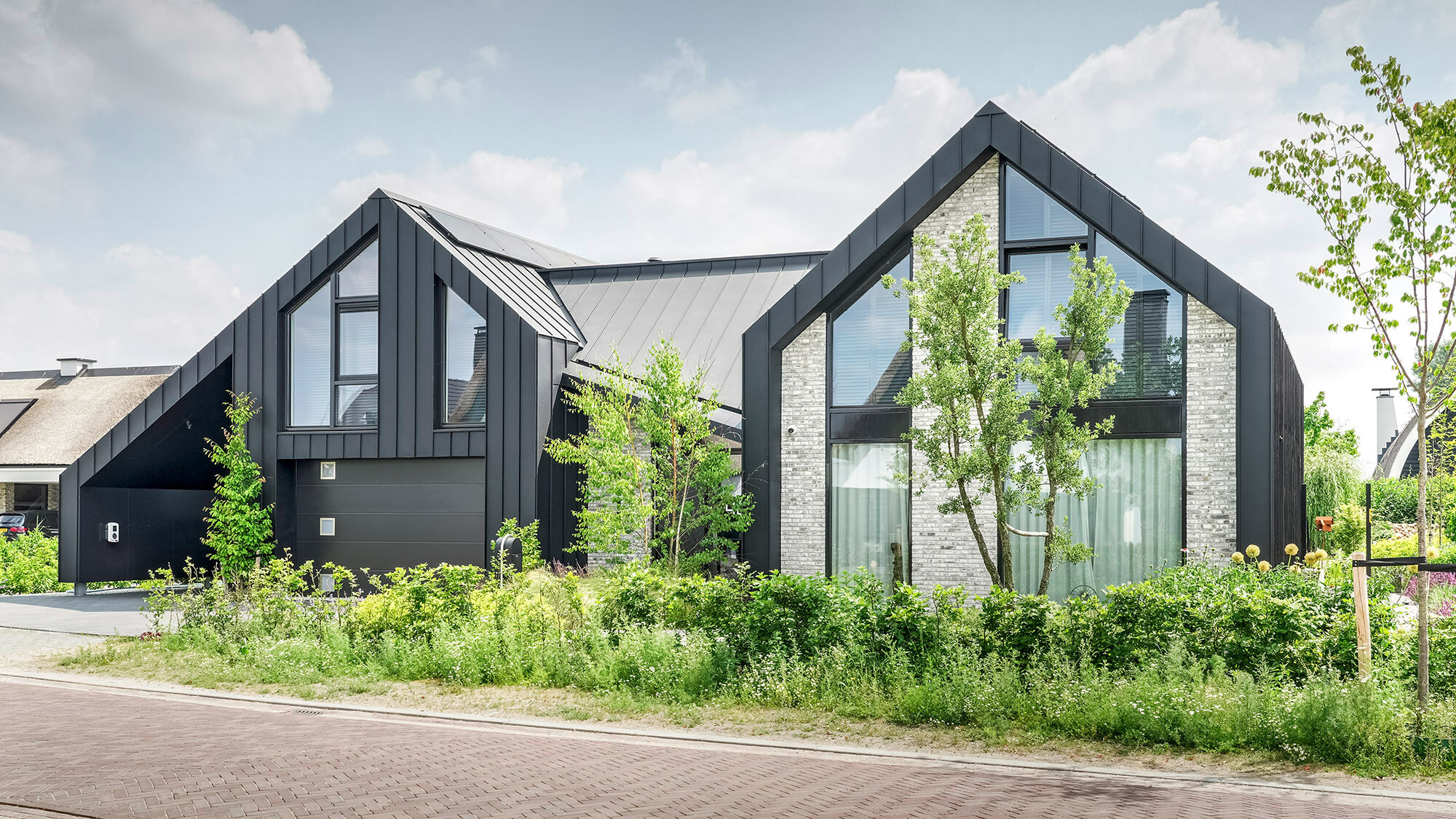 View of a modern detached house with pointed gable roofs made of black Prefalz material, located in a quiet street. The house combines a dark façade with light-coloured brick accents and large areas of glazing. Young trees and a naturally landscaped front garden frame the house and blend into the peaceful neighbourhood.