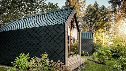 Ein minimalistisches Tiny House des Secret Koi Garden am Natterer See steht eingebettet im Grünen und wird von warmem Abendlicht umrahmt. Dach und Fassade sind vollständig mit kleinteiligen PREFA Dach‑ und Wandrauten aus Aluminium in P.10 Anthrazit verkleidet, deren präzise, fischhautartige Struktur markante Akzente setzt. Das langlebige, witterungsbeständige PREFA Aluminium schützt die Architektur zuverlässig und harmoniert perfekt mit Holz, Glas und der umliegenden Natur.