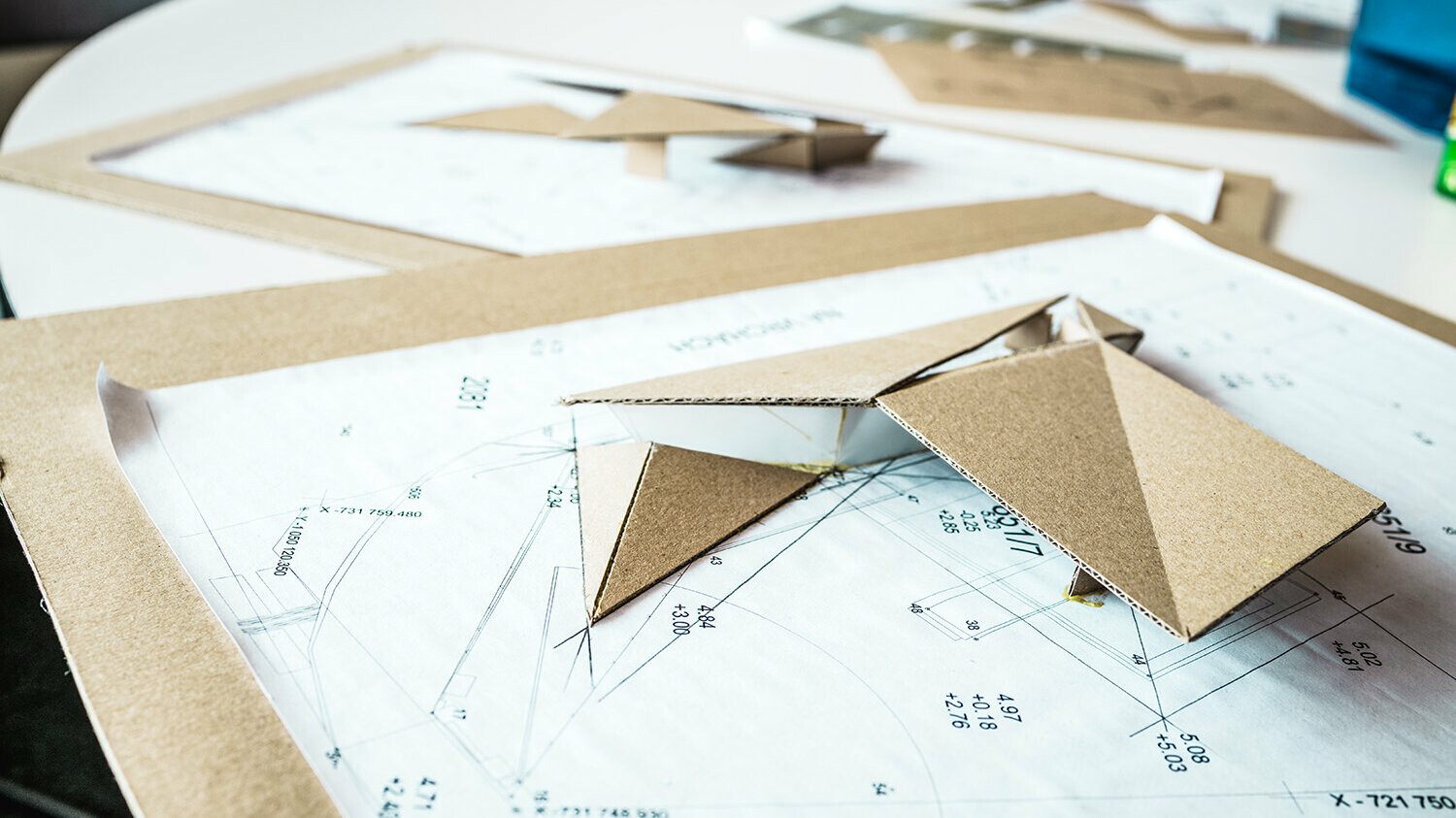 Photo of plans of the filling station with a blurred background, a cardboard model of the folded roof rests on one of the plans.