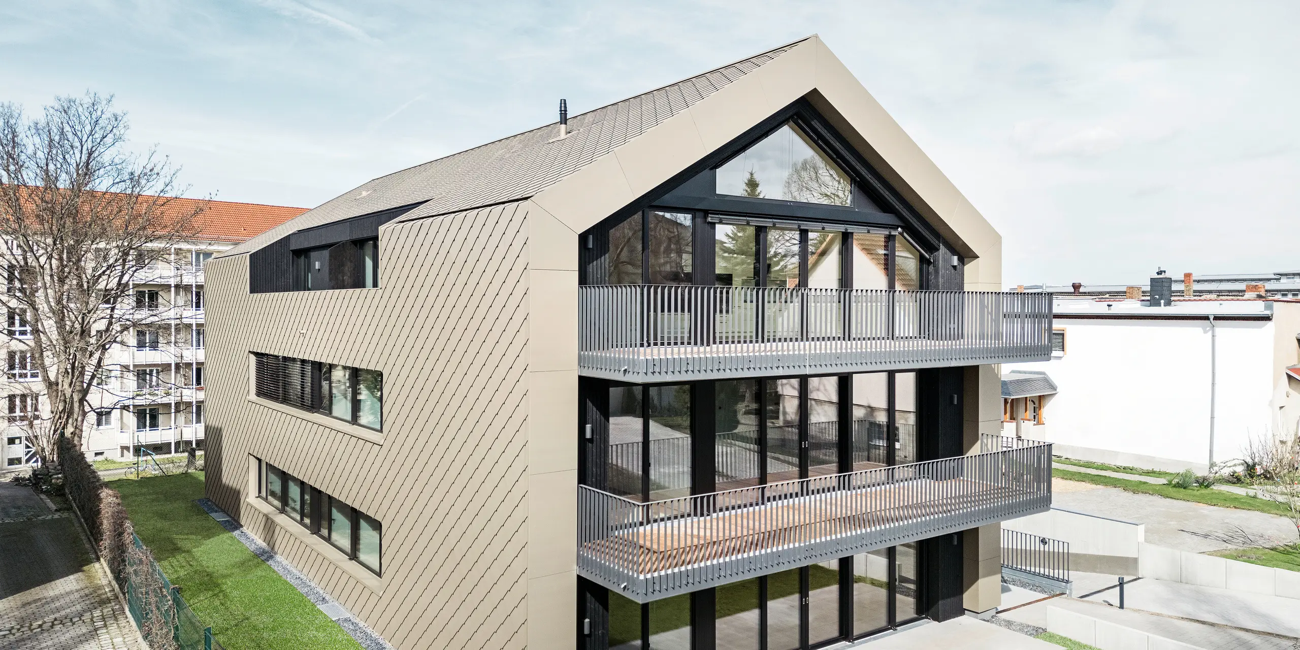 Gartenansicht eines modernen Mehrfamilienhauses in der Chemnitzer Straße in Dresden. Der gepflegte Garten liegt vor einer eindrucksvollen Architektur. Die Fassade und das Dach des Gebäudes sind mit der langlebigen PREFA Aluminiumraute 29 × 29 in Bronze verkleidet, die dem Bauwerk eine elegante und zeitgemäße Optik verleiht. Das Bild zeigt die großzügigen Balkone, die großen Fensterfronten und die harmonische Einbettung des Gebäudes in die Umgebung.
