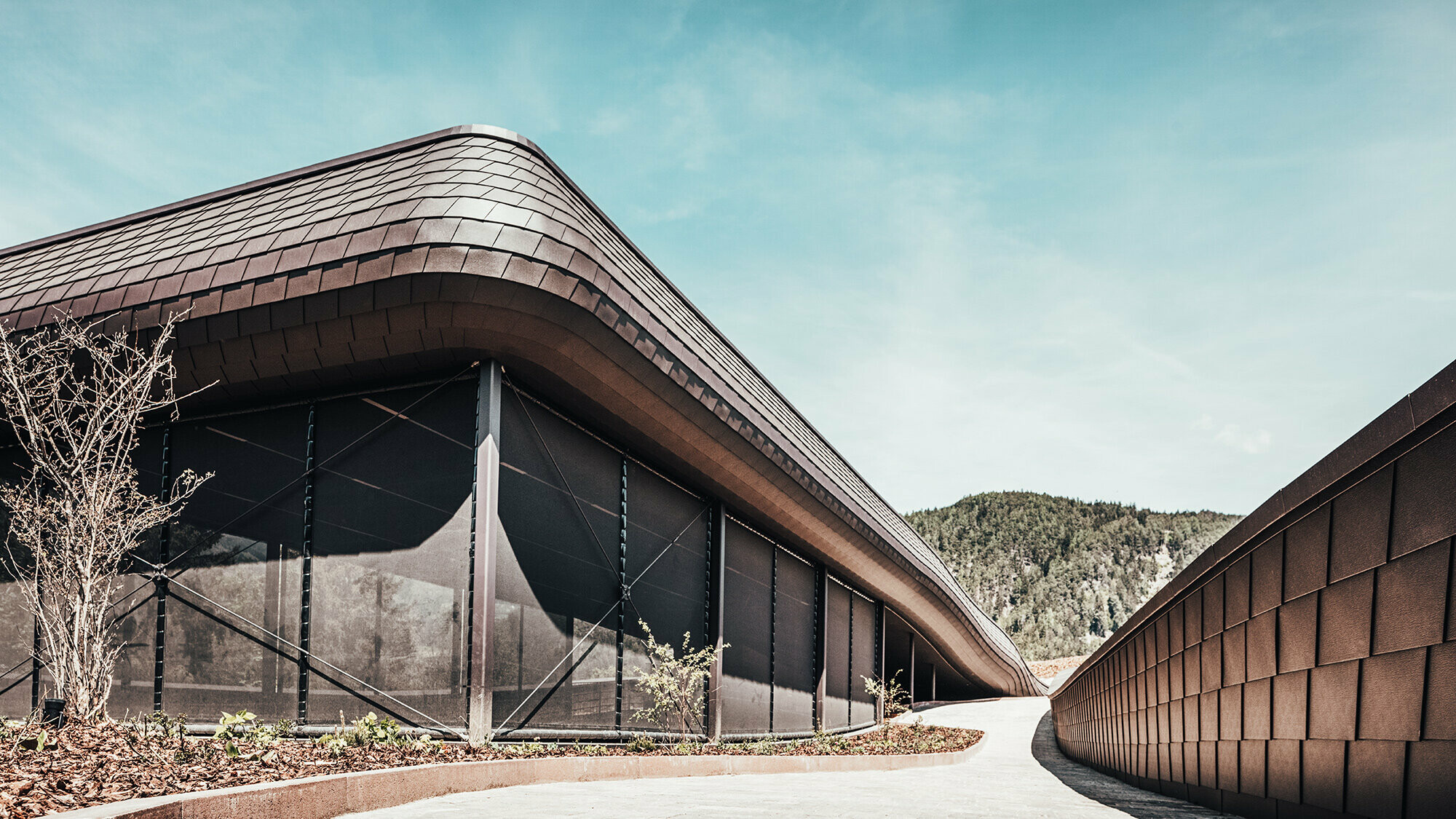 Side view of the exterior of the hotel in South Tyrol. The PREFA façade shingles can be seen on every surface. The hilly, wooded landscape can be seen in the background.