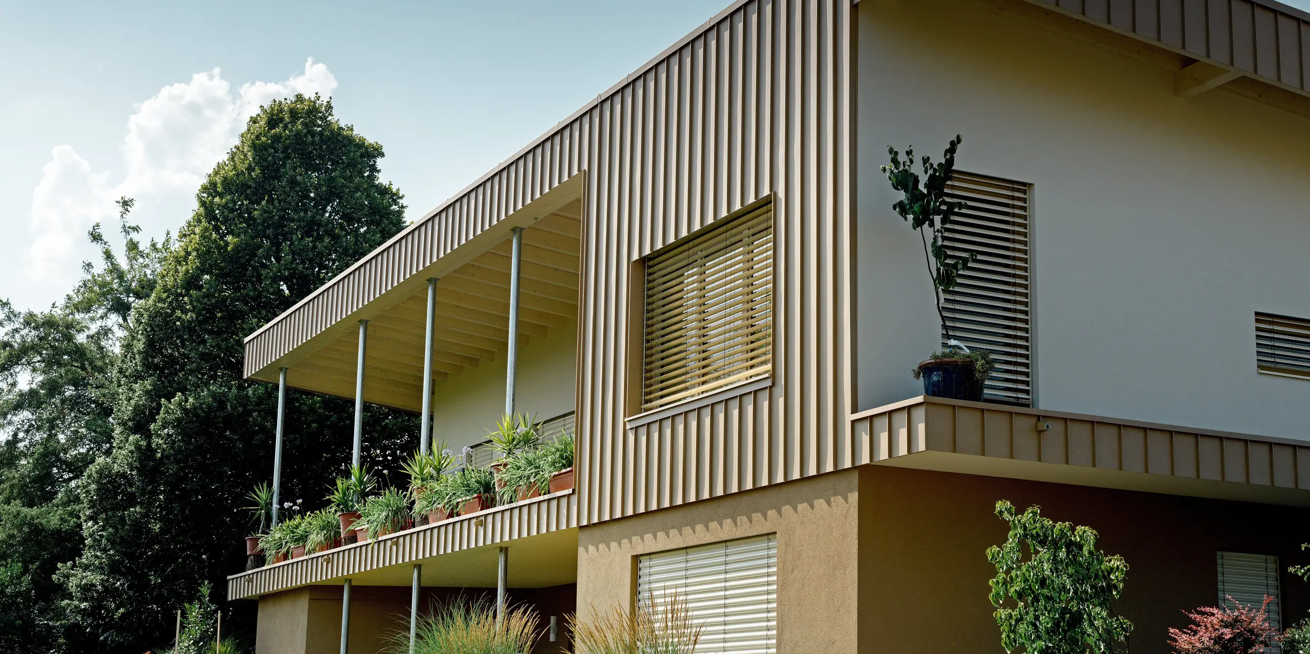 Perspektivische Ansicht des Einfamilienhauses in Schiefling am See mit markanter PREFALZ Fassade aus Aluminium in P.10 Sandbraun. Die prägnanten Falzprofile rahmen die Architektur eindrucksvoll ein und lenken den Blick auf die großzügige Terrasse mit üppiger Begrünung. Die langlebige PREFA Aluminiumfassade überzeugt durch zeitlose Eleganz, Wartungsarmut und hohe Witterungsbeständigkeit