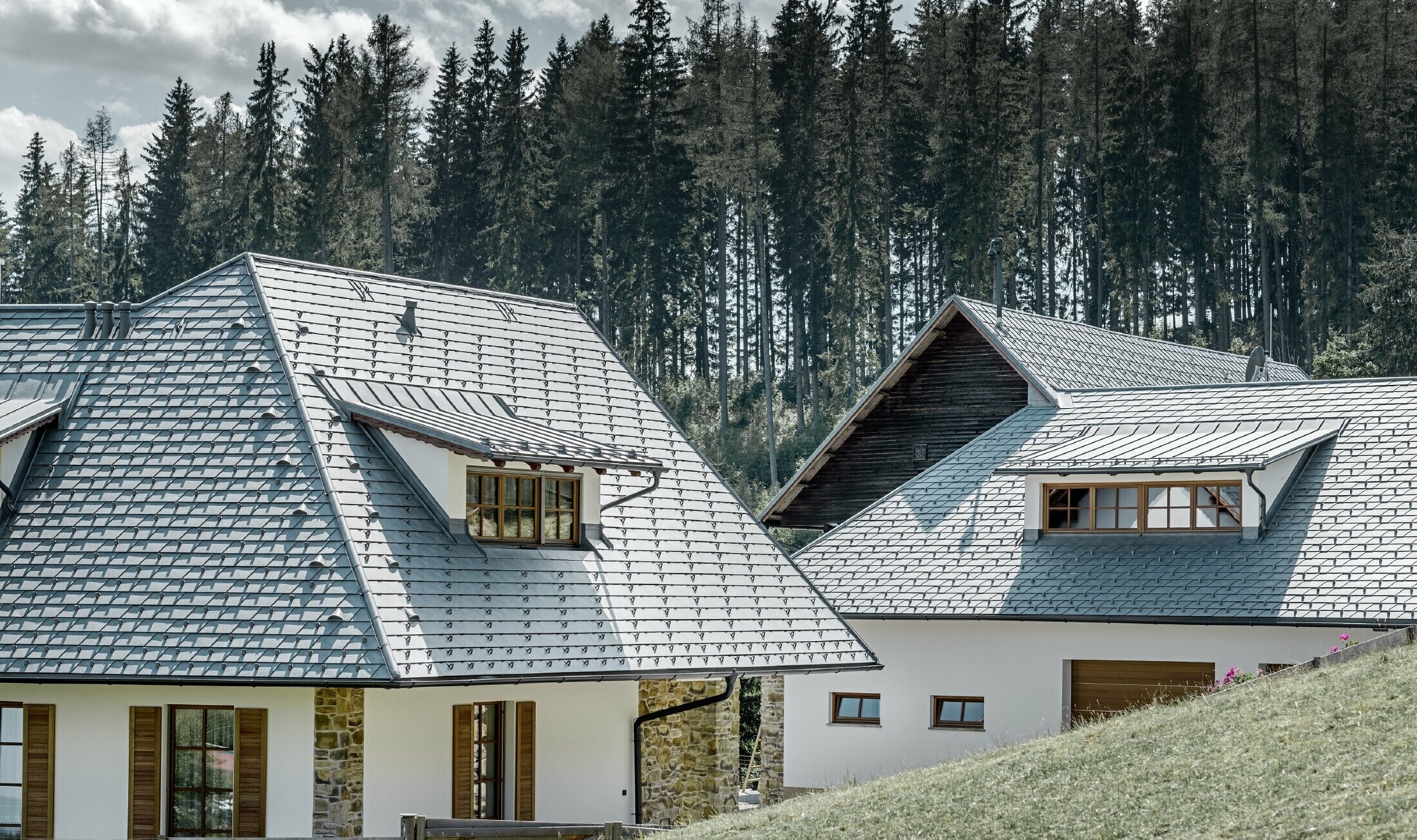 Rear view of the detached house on a slope; the roof was clad in aluminium roof shingles in stone grey, the eyebrow dormers with standing seam are also in stone grey. A forest can be seen in the background.