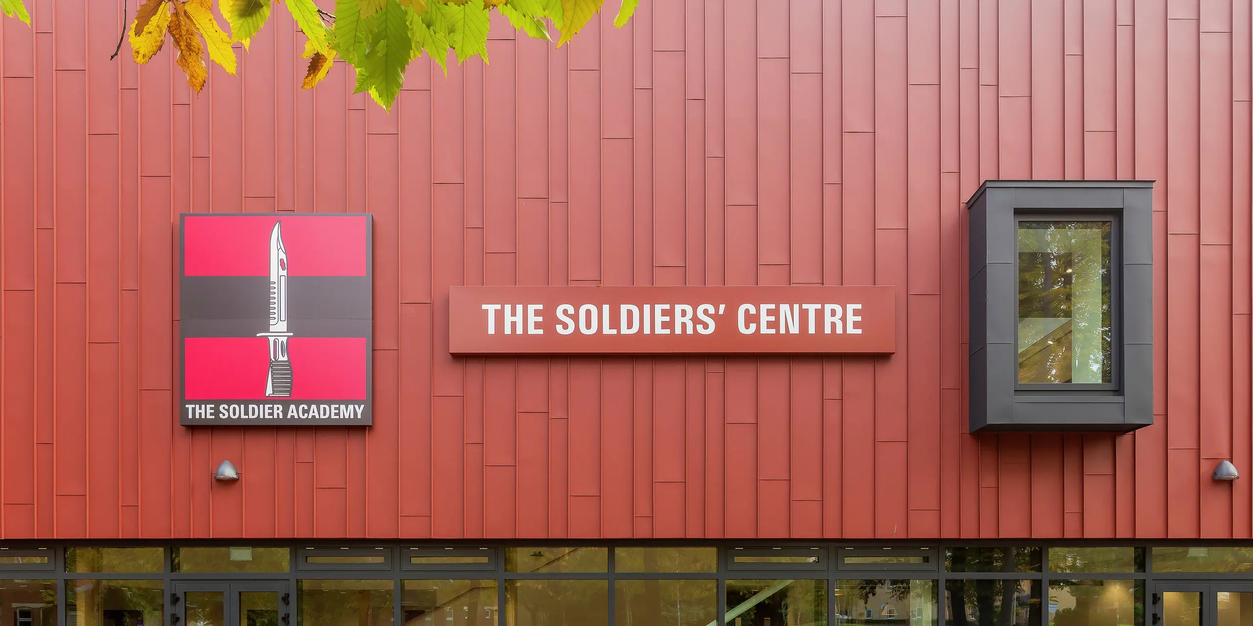 Purpose-built army welfare facility with PREFALZ façade: the precisely manufactured standing seam roof in P.10 oxide red gives the building a striking appearance. The entrance area is impressive with its generous glass fronts, providing brightness and transparency. The 'Soldier Academy' logo and the eye-catching building sign provide a clear visual focus. The window frames in P.10 anthracite round off the design harmoniously. High-quality PREFA aluminium products for durable and aesthetic architecture.