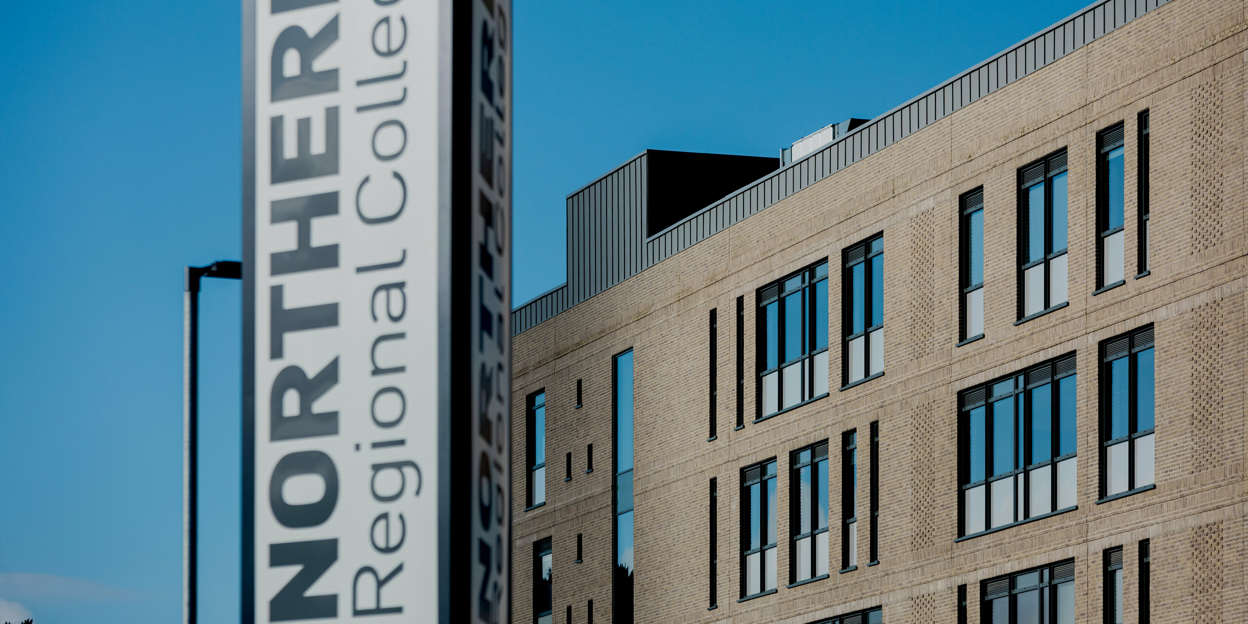 A vertical sign reading “Northern Regional College” dominates the foreground, with the brick campus buildings of Ballymena visible behind it against a clear blue sky. Along the roofline, PREFALZ standing seam aluminium panels in patina grey form a crisp horizontal edge that defines the architecture. Narrow, evenly spaced windows create a strong façade rhythm. PREFA aluminium enhances the building with durability, low maintenance and a refined, contemporary appearance suited for modern educational architecture.