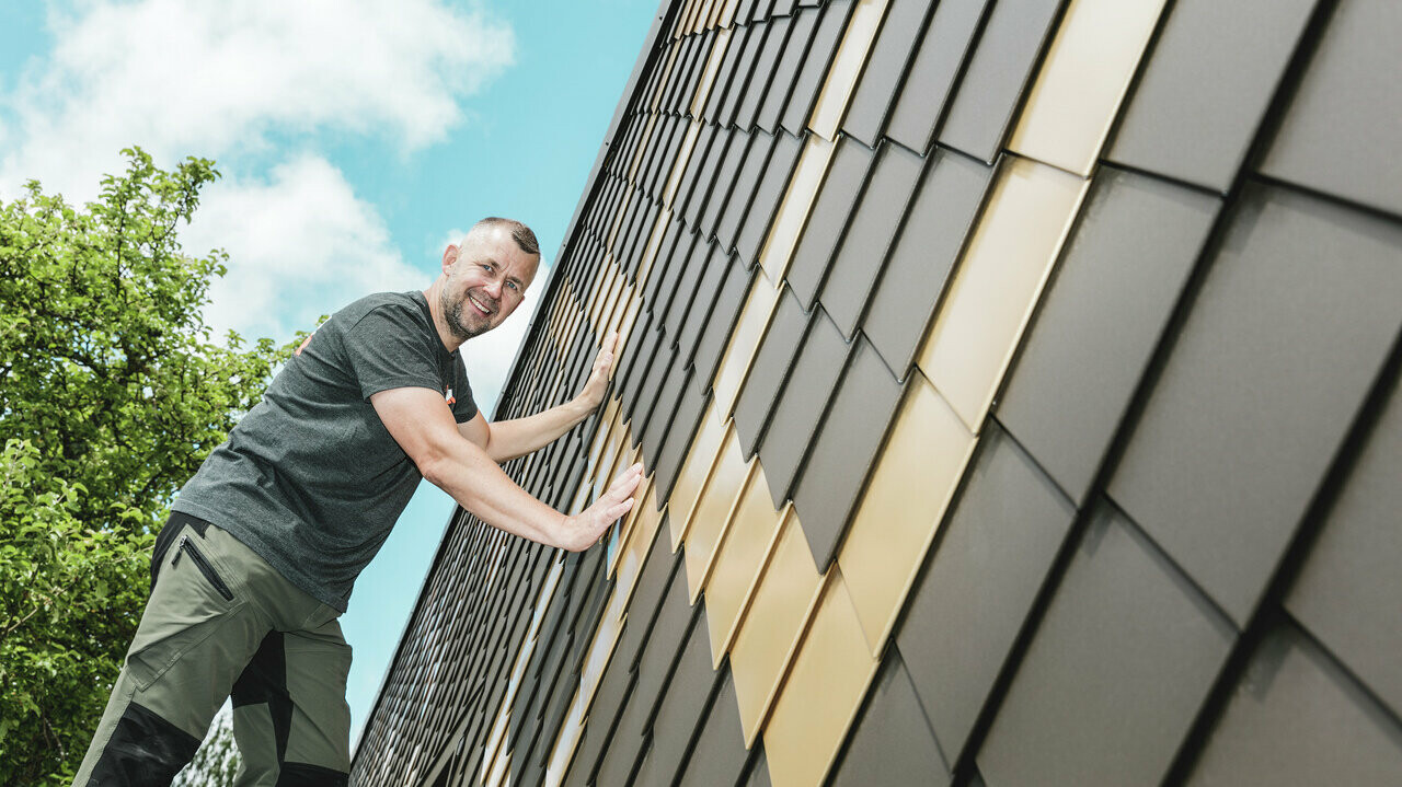 PREFA tradesman in a grey T-shirt with a PREFA rhomboid tile façade in brown and Mayan gold.