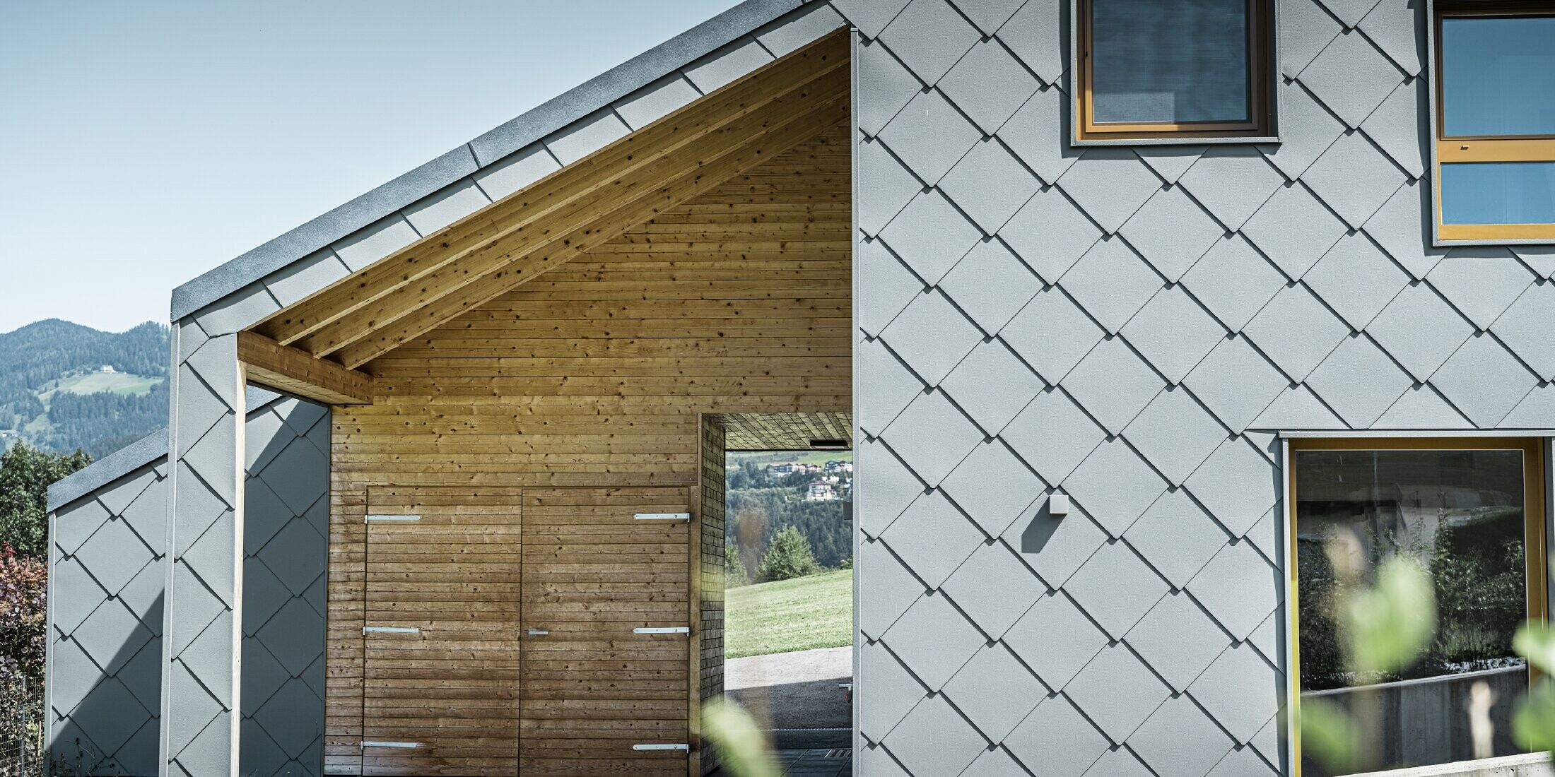 The covered entrance area is made with horizontal wood panelling, the rest of the façade is clad with the large PREFA aluminium rhomboid façade tiles in light grey